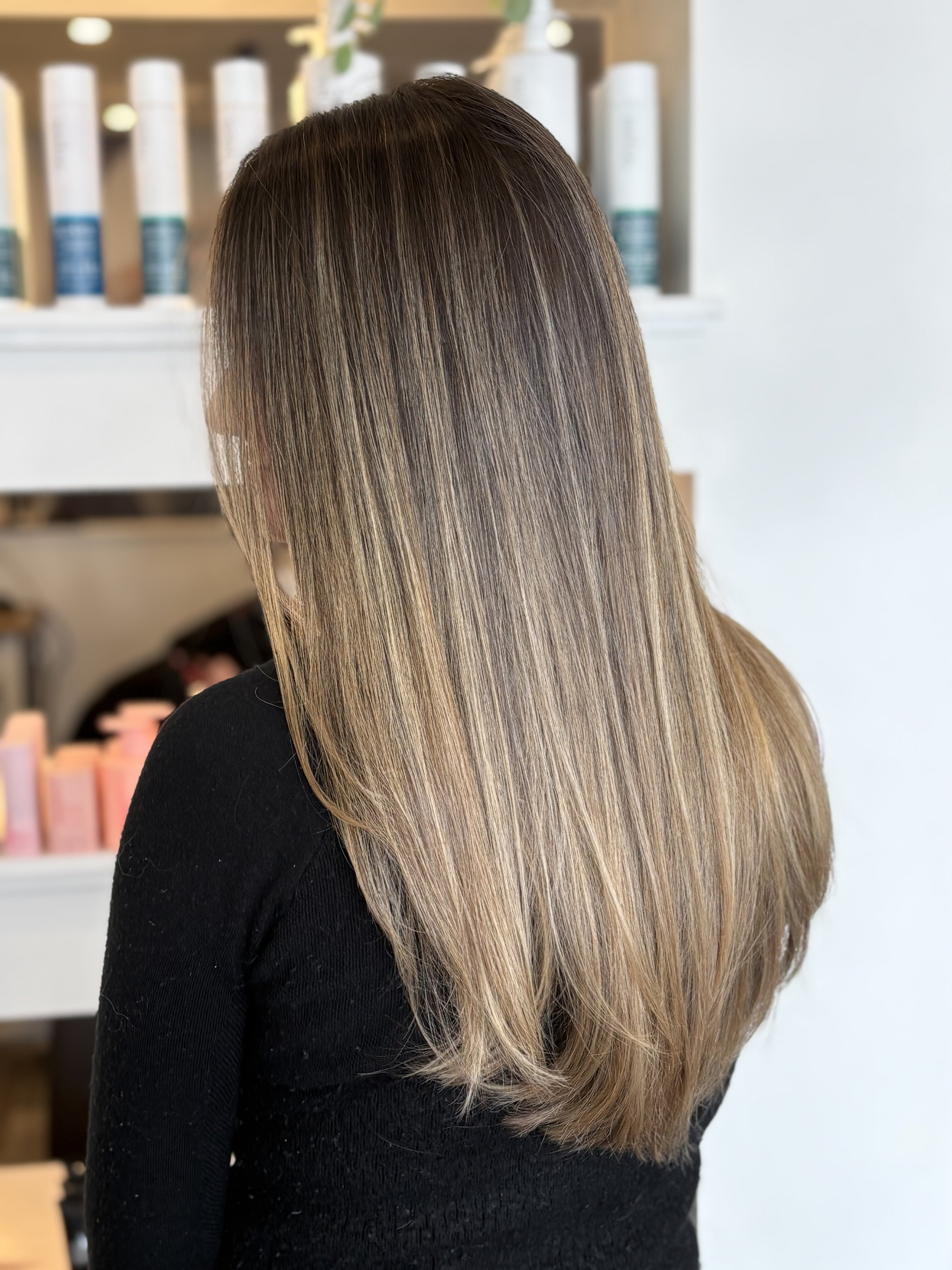 A person with straight, long, shiny hair with highlights, seen from behind, wearing a black top. The background includes shelves with hair products.