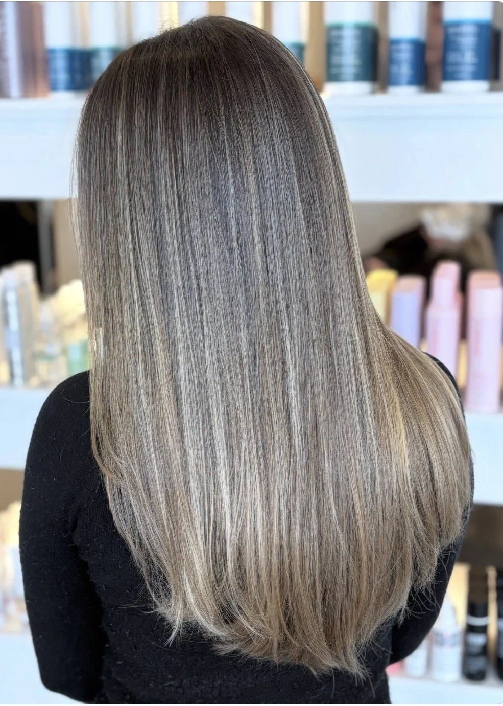 Back view of a woman with long, straight, shiny blonde hair, standing in front of shelves with hair care products.