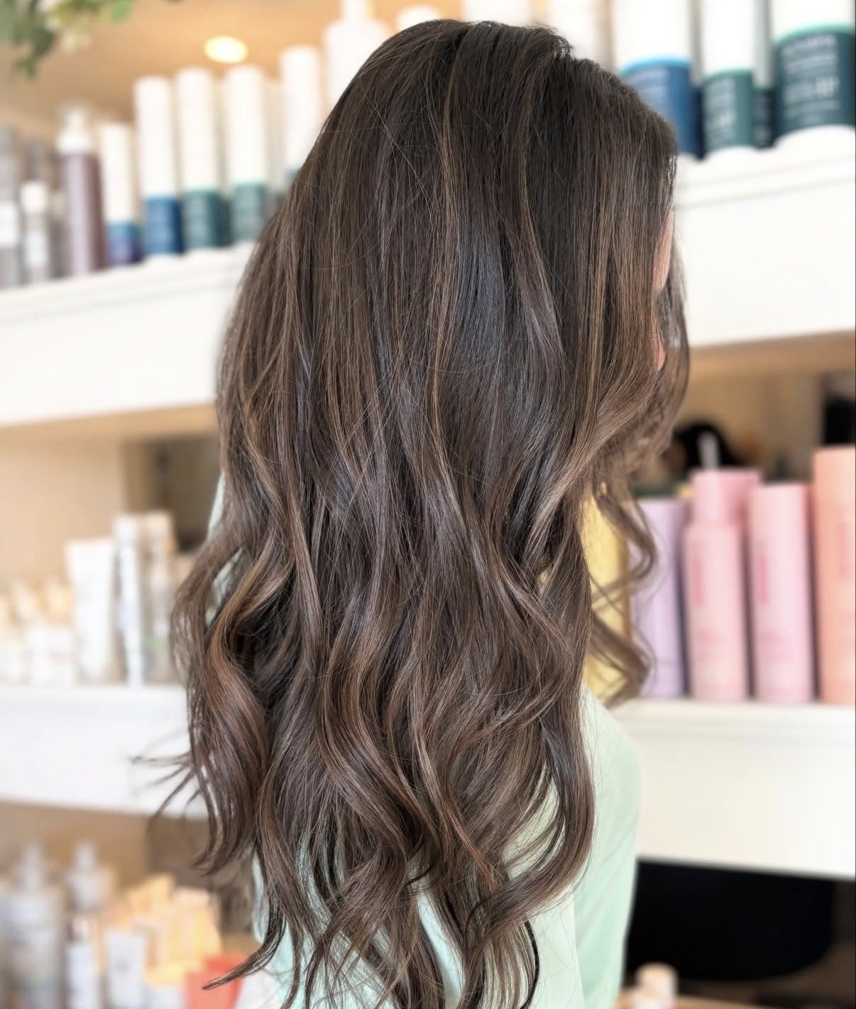 A woman with long, wavy brunette hair standing in front of shelves with hair care products.