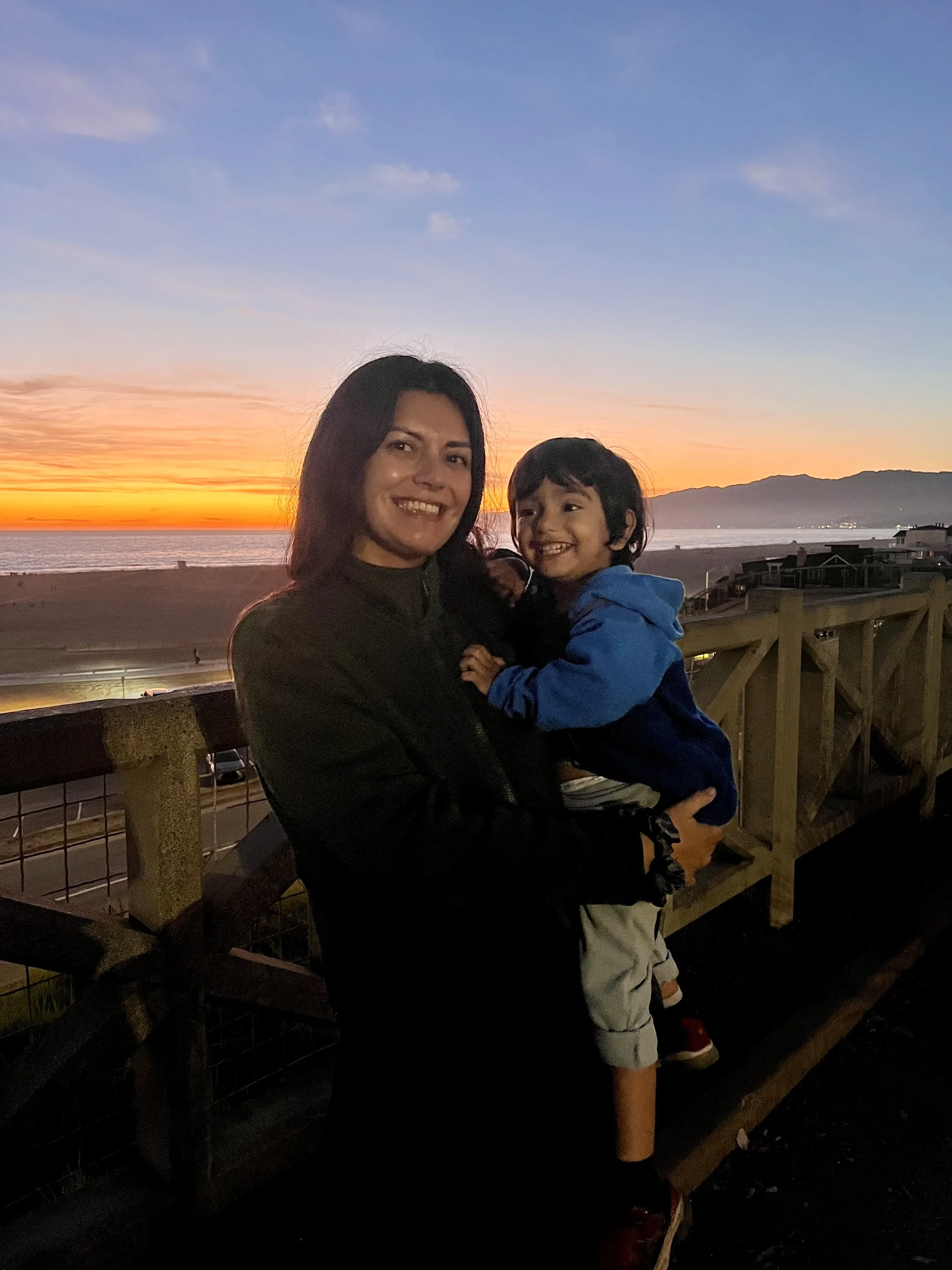 A woman and a young boy smiling and hugging on a wooden platform near the ocean at sunset.