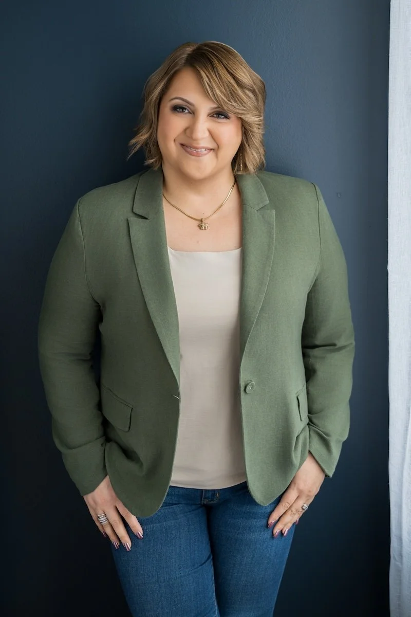 Sara smiles directly into the camera. She wears a green blazer and is posing in front of a blue wall.