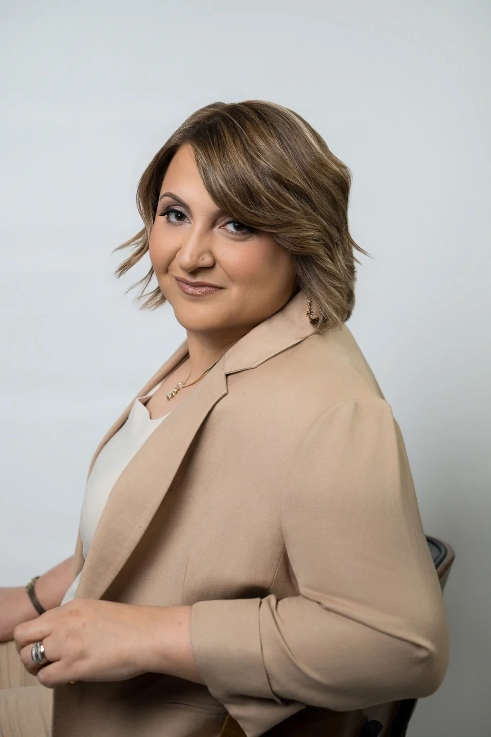Sara sits slightly turned toward the camera, she wears a tan jacket with her arm bent