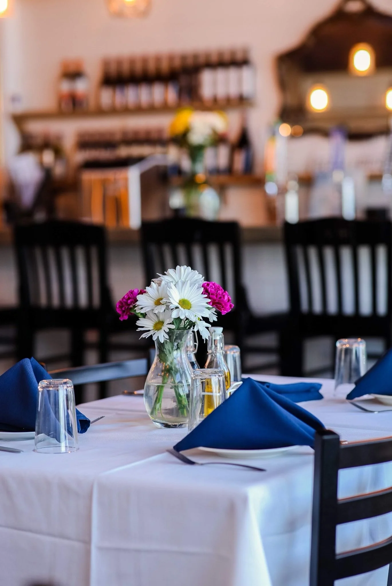 A table set for a formal event with a white tablecloth, a glass vase with white and pink flowers, blue napkins, and glassware. In the background, a dining area with chairs, a wall with shelves, and warm lighting.