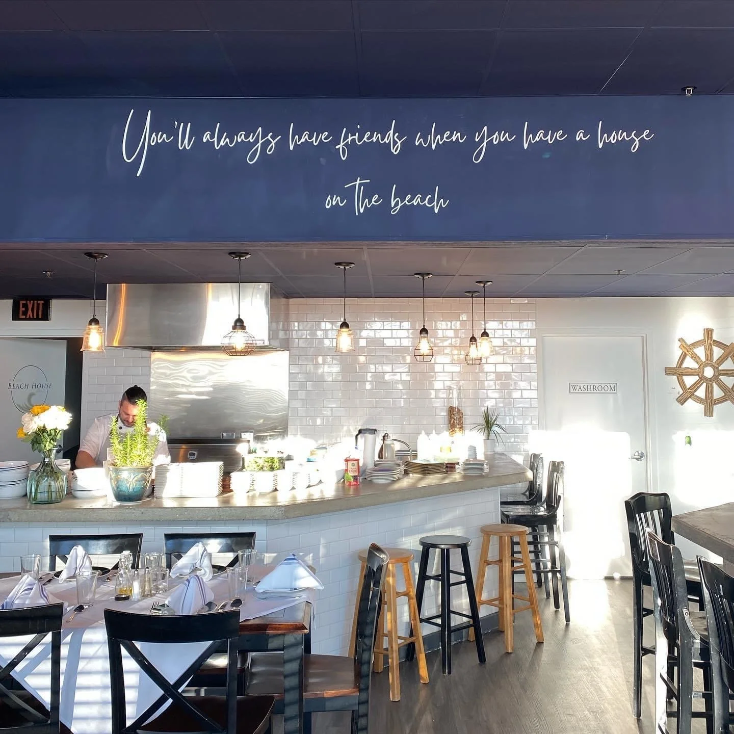Interior view of a modern restaurant with a white-tiled bar, hanging light fixtures, a sign on the wall that says 'Washroom', and a large blue sign with a quote about friendship. There are tables set with white napkins and glasses, and a few barstool