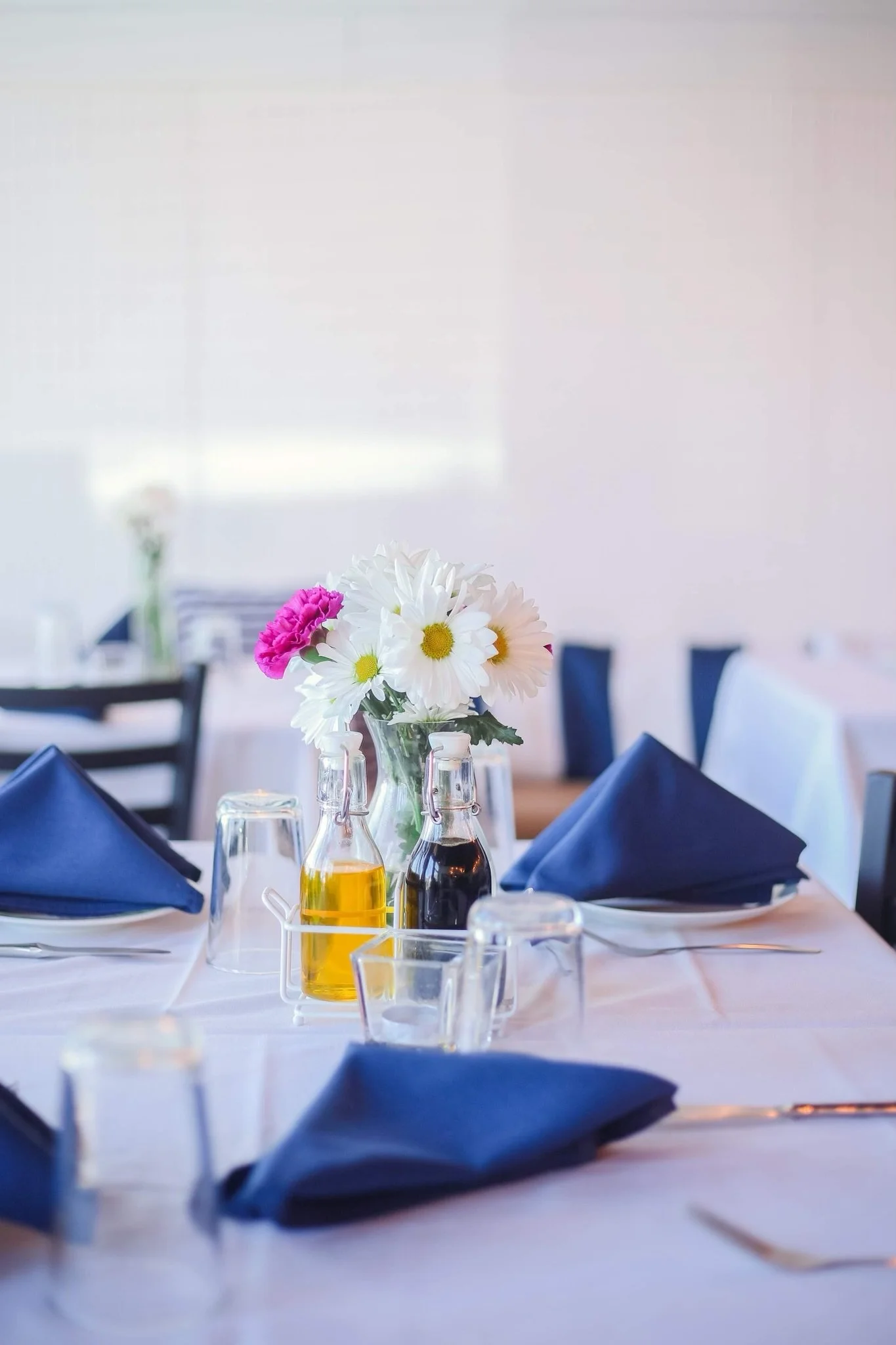 A dining table set with a small bouquet of white daisies and pink carnations, blue napkins, glass bottles of oil and vinegar, and empty wine glasses in a bright, minimalist restaurant or event space.