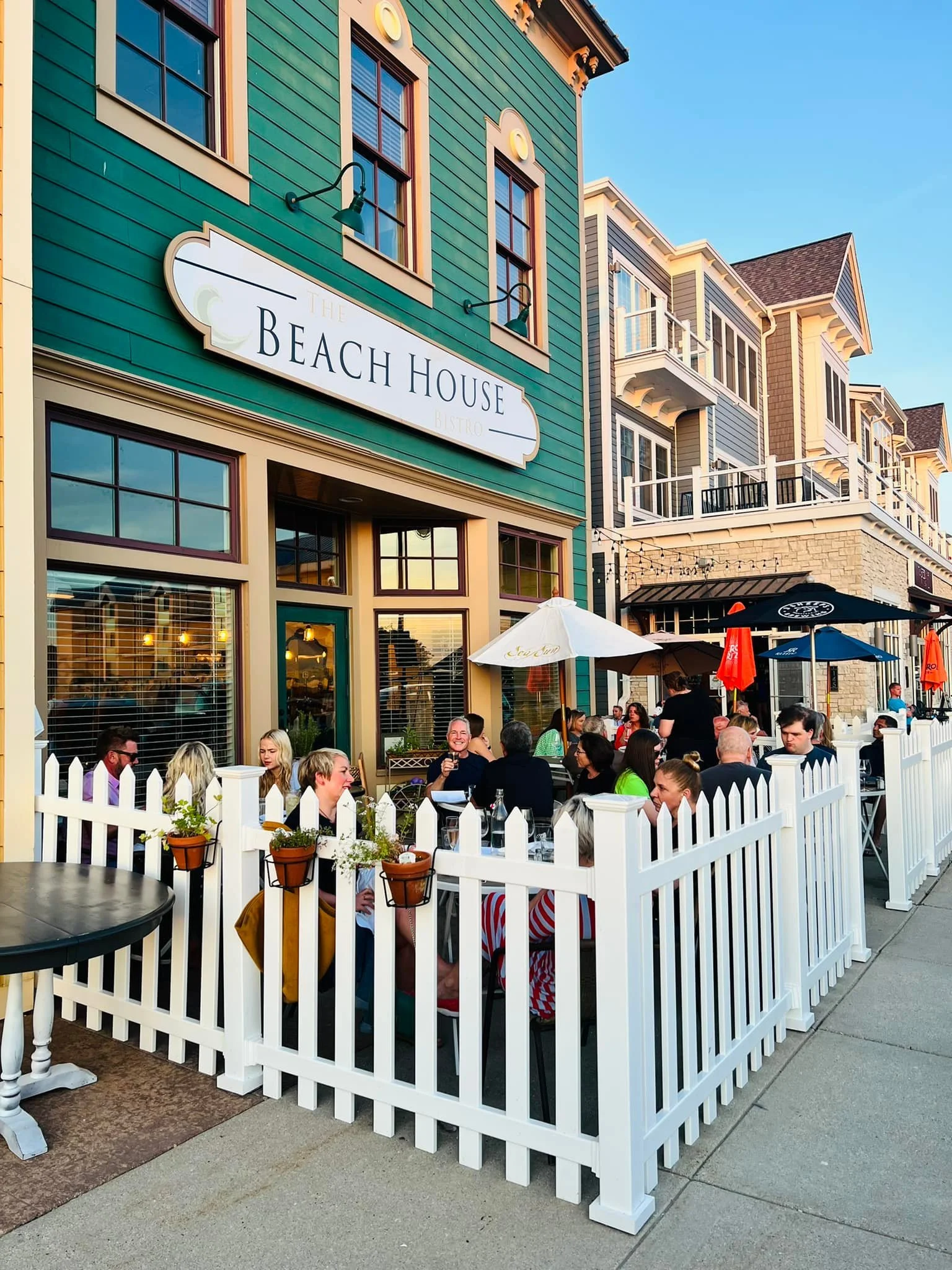 People dining at outdoor seating area of The Beach House Bistro, with building featuring colorful Victorian-style architecture in the background.