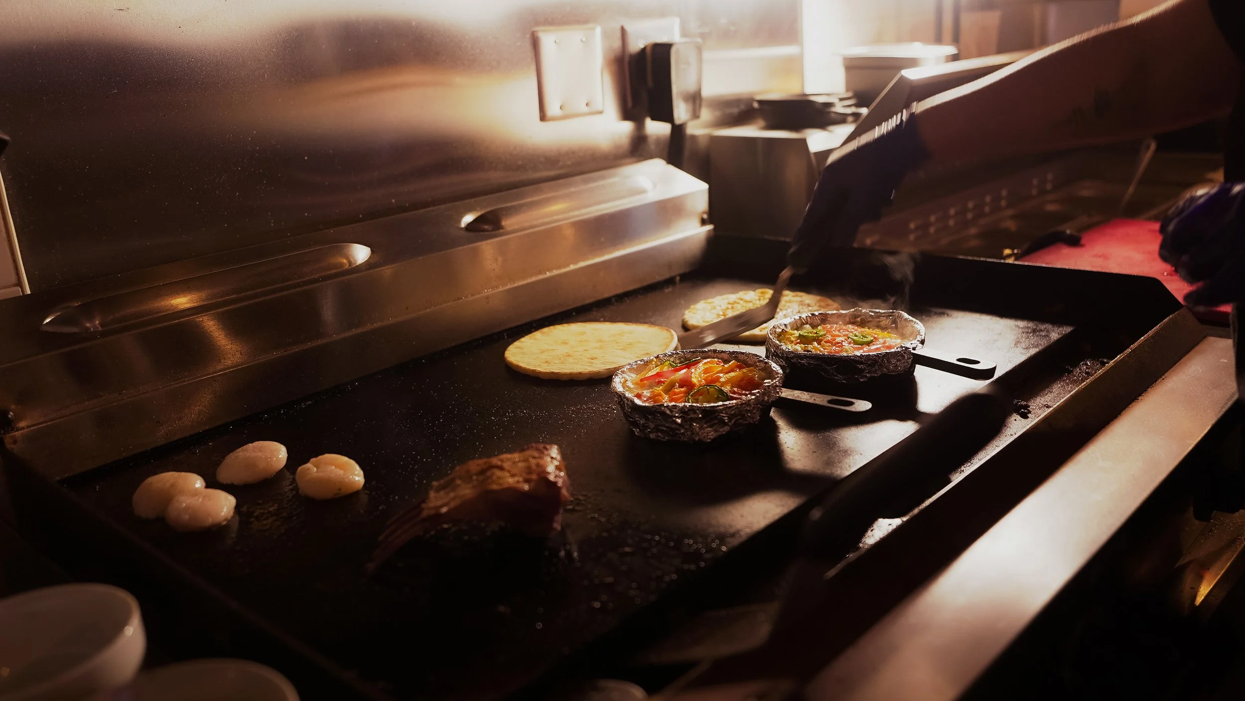 Food being cooked on a flat grill, including tortillas, small cast iron skillets with toppings, and pieces of meat. A person's hand is using tongs to handle the food.