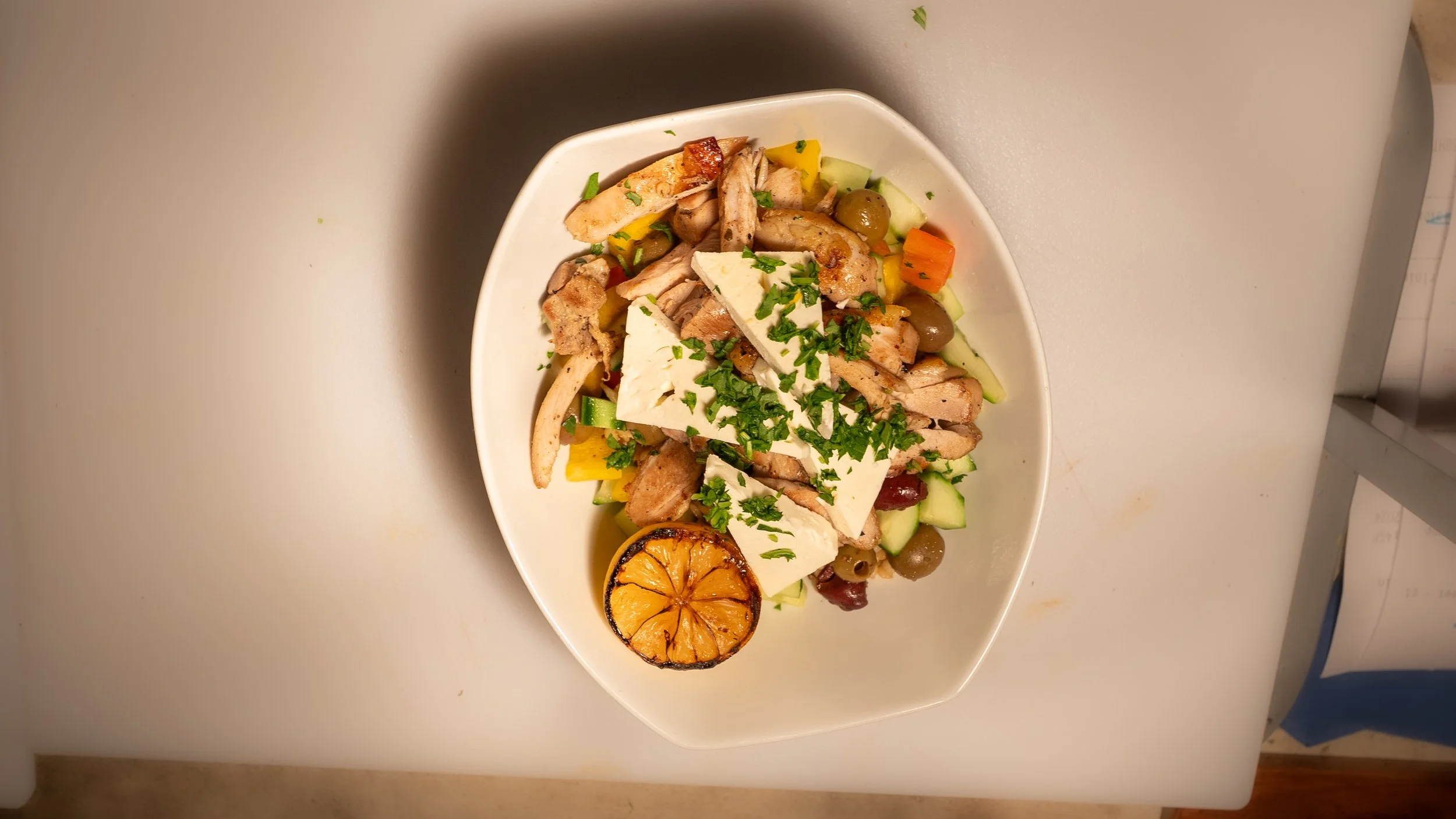 A bowl of salad with grilled chicken, cheese, olives, and vegetables, garnished with herbs and a grilled lemon half.