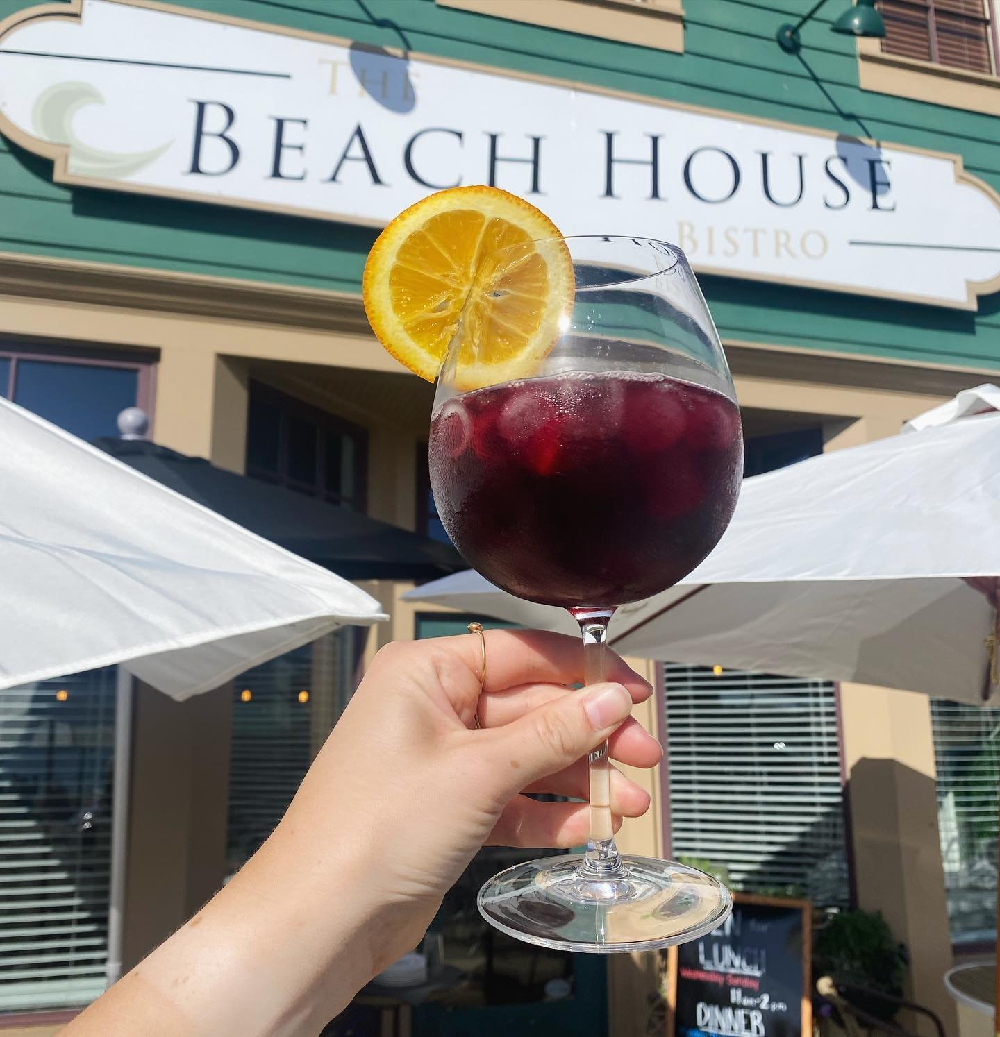 A hand holding a glass of red wine with ice and a lemon slice garnish, with a restaurant called 'The Beach House Bistro' in the background, outdoor patio with umbrellas.