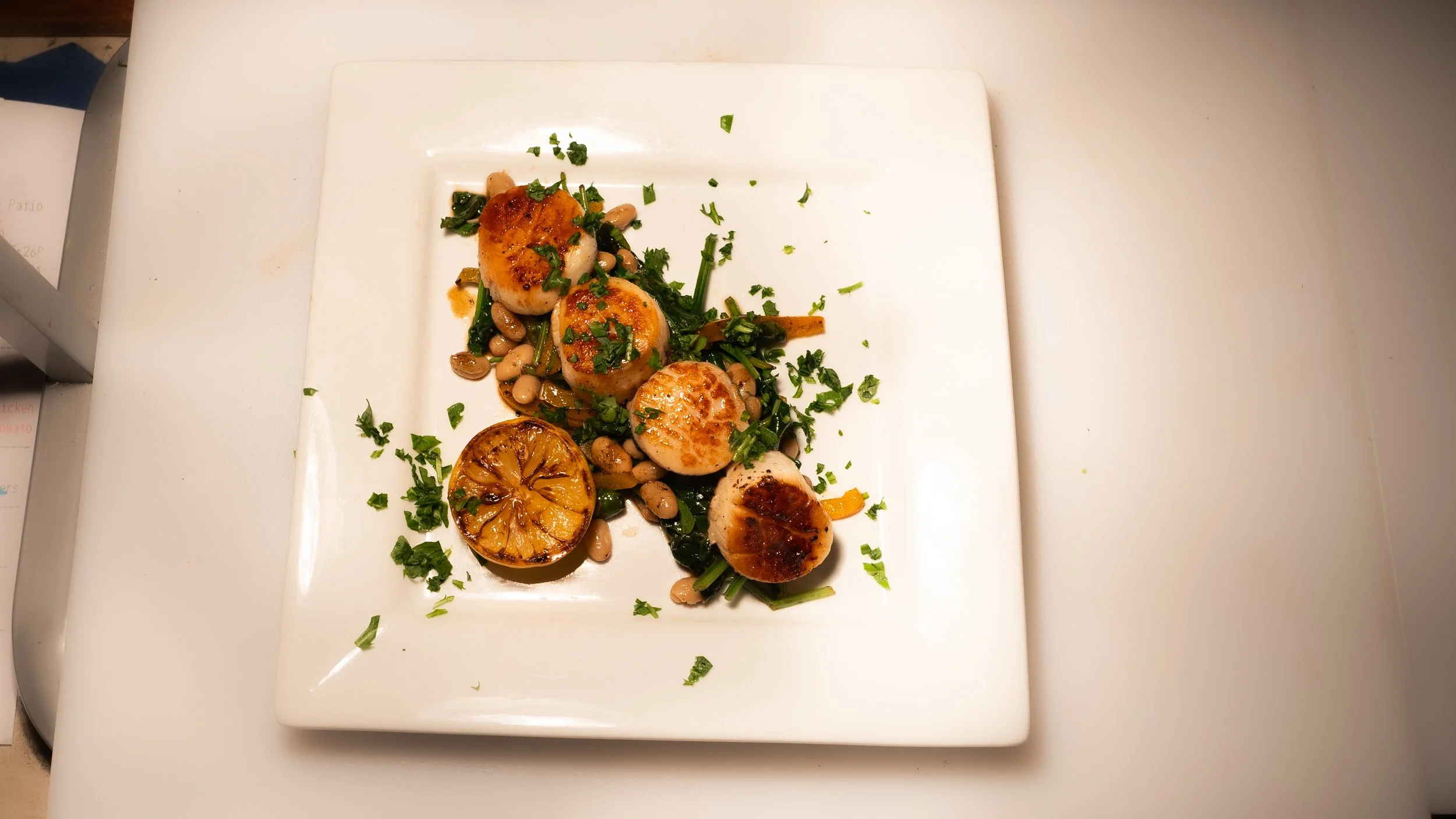 Seared scallops garnished with chopped herbs, served with grilled lemon, white beans, and sautéed greens on a white square plate.