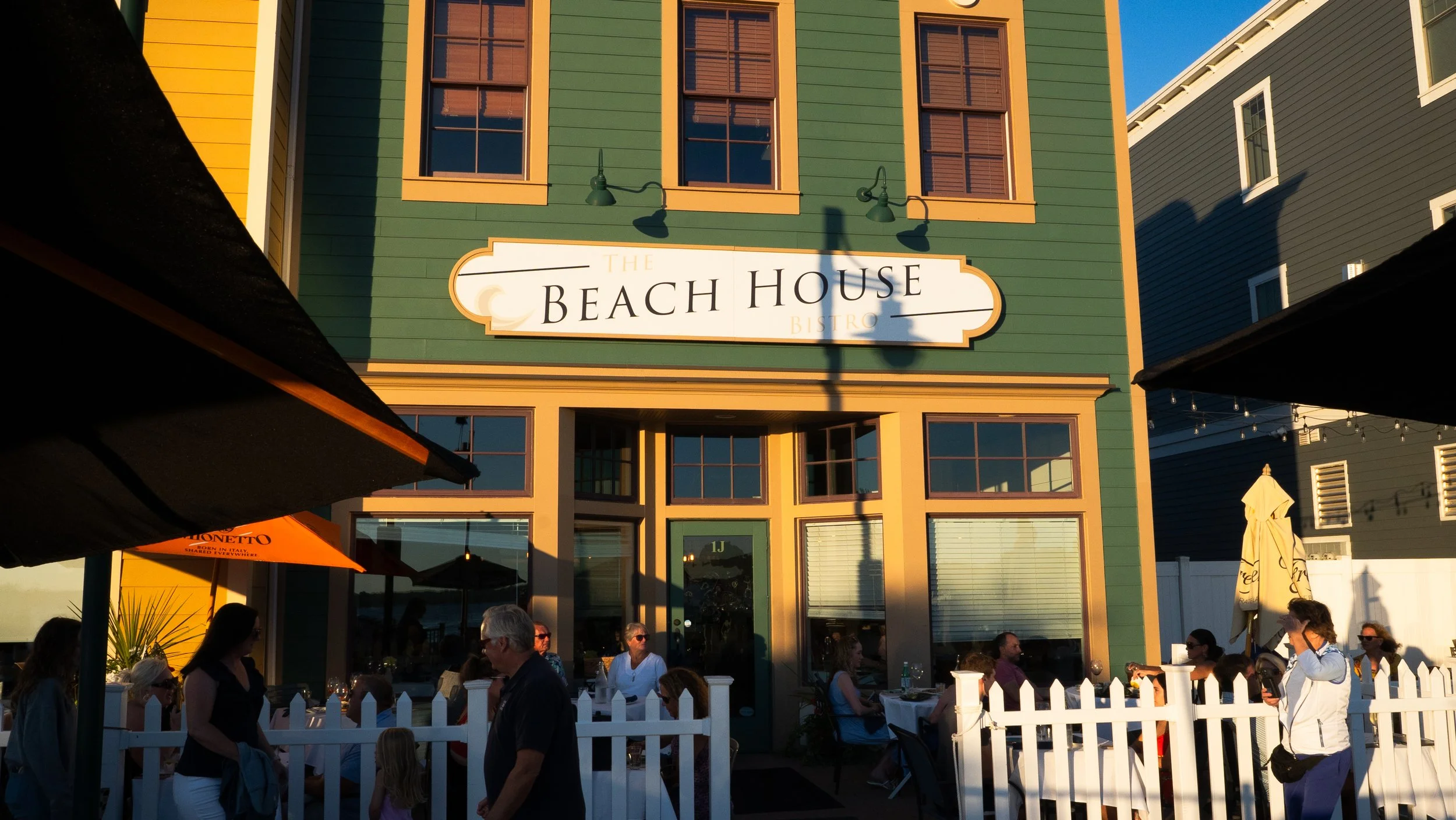 People dining outdoors at The Beach House Bistro, a green multi-story building with large windows and a white sign, with umbrellas and string lights visible.
