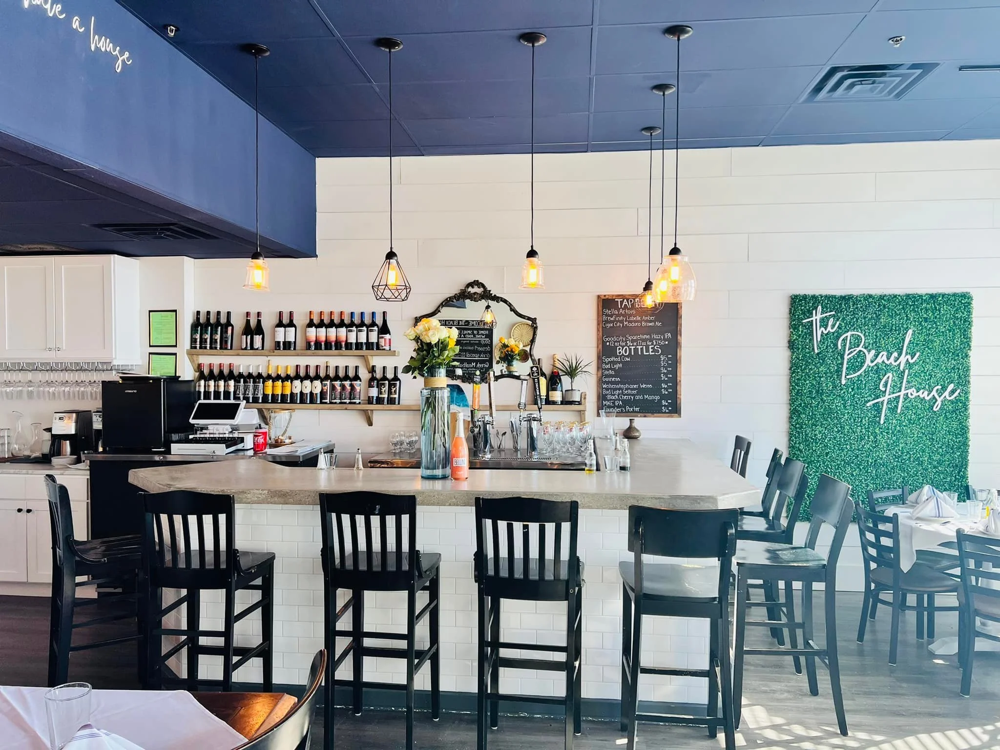 Interior of a modern restaurant or café with a bar area, black chairs around tables, blue ceiling with pendant lights, wine bottles on shelves, a chalkboard menu, and decorative elements including a green textured wall with 'The Beach House' written 
