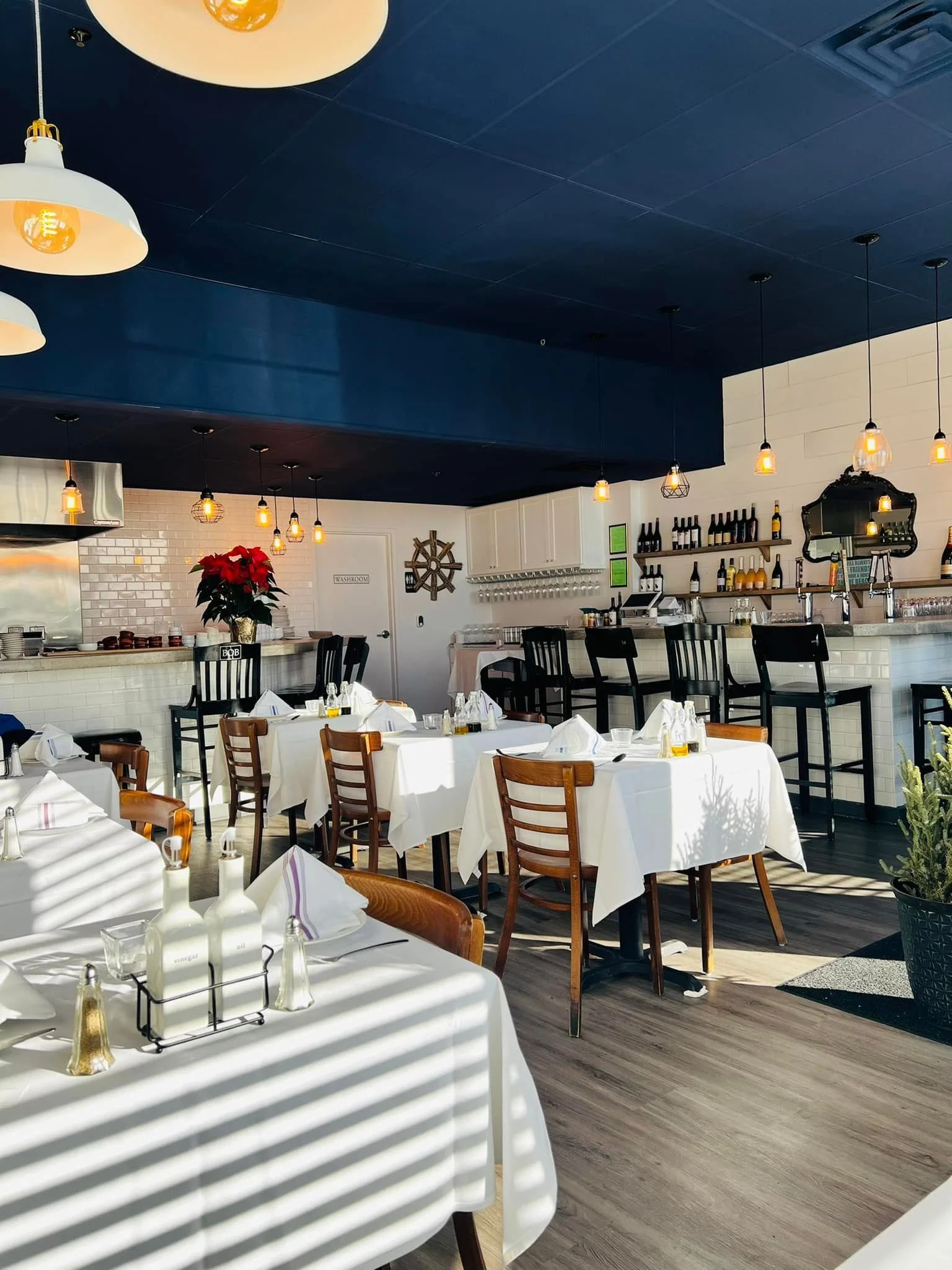 Interior of a restaurant with white tablecloths, wooden chairs, and modern hanging light fixtures. There is a blue ceiling and a white brick wall decorated with nautical-themed items and wine bottles on shelves.