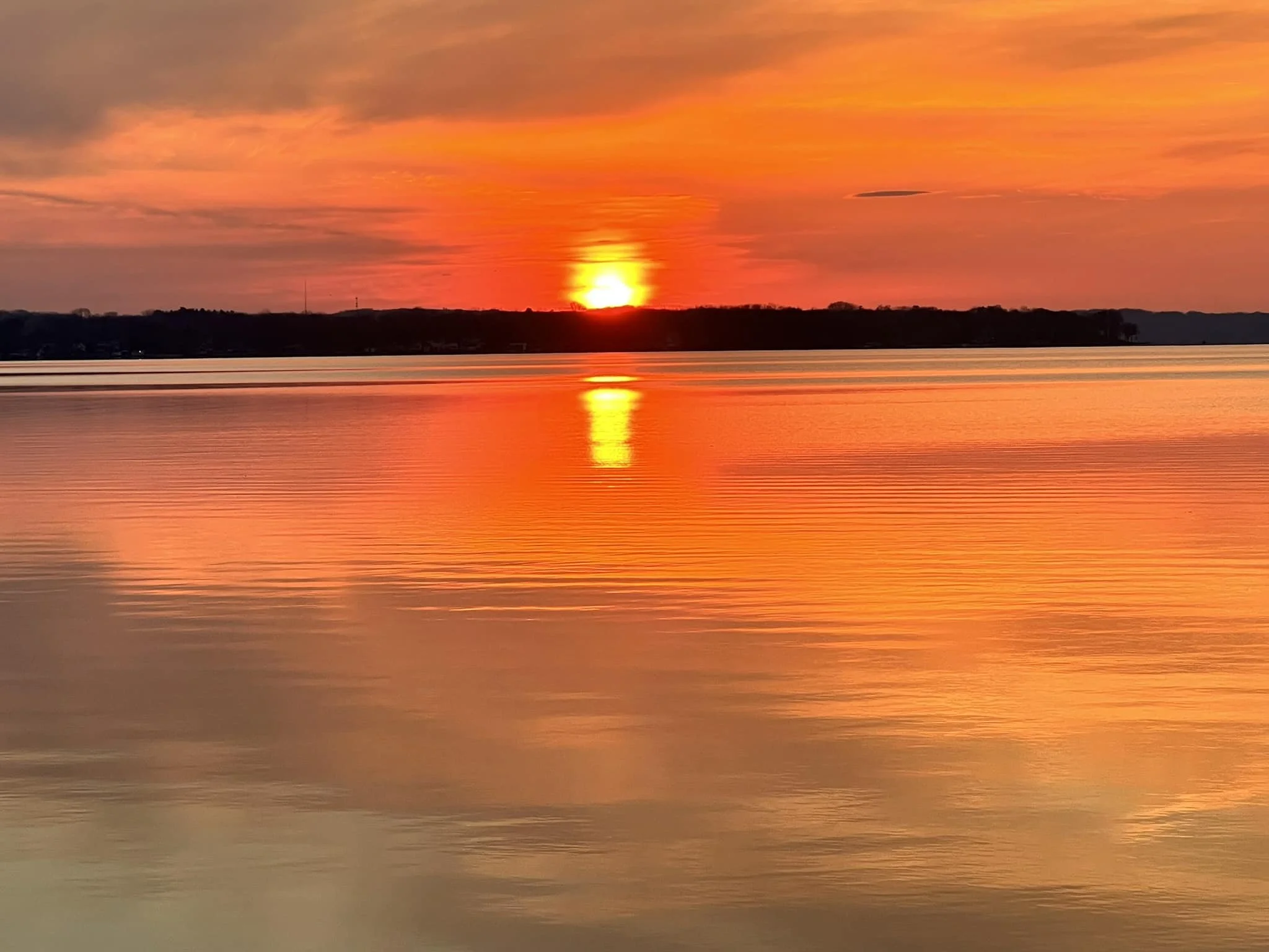 A vibrant sunset over a calm body of water with a silhouette of land and trees in the distance.