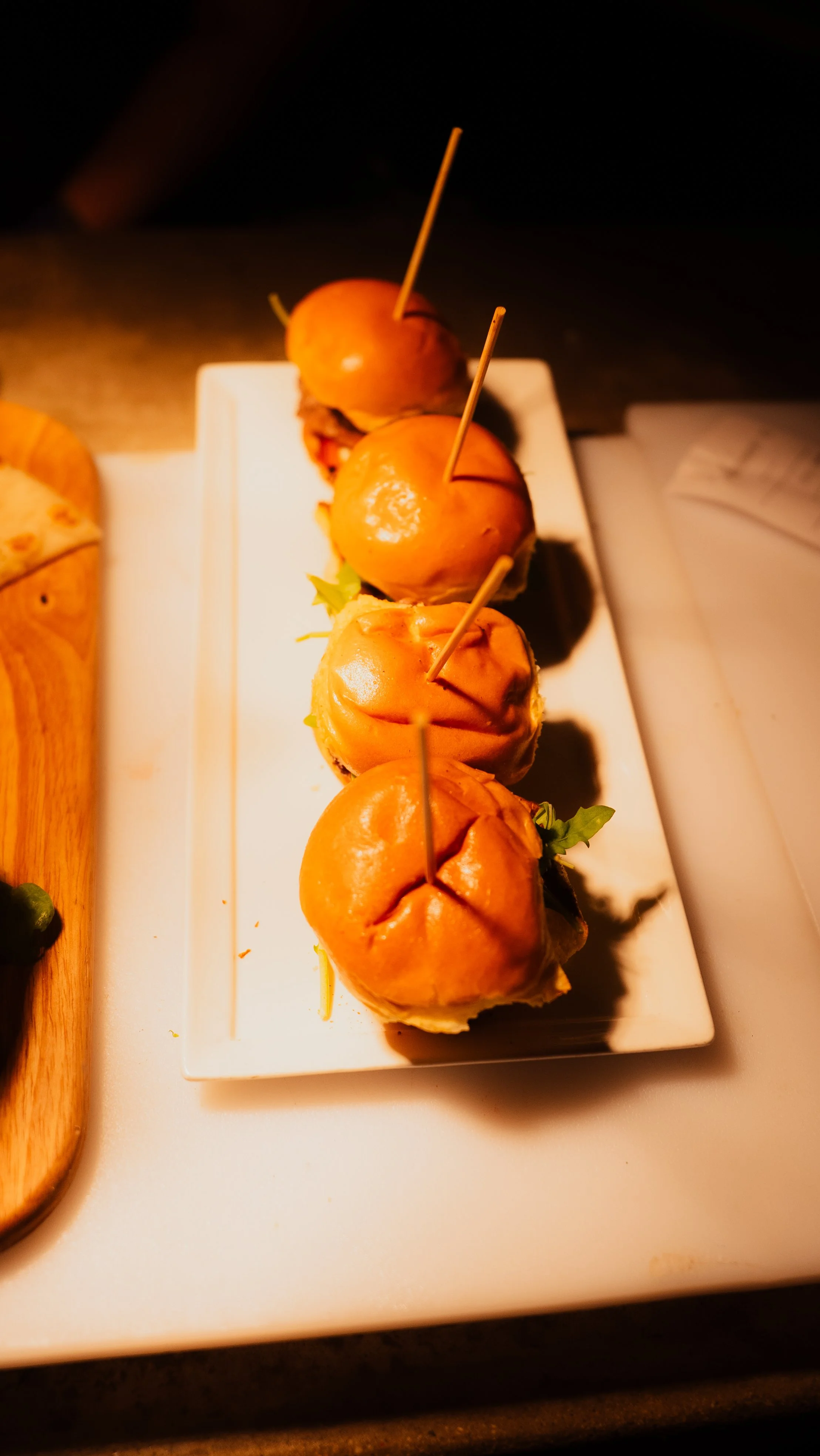 Close-up of four small burgers with buns, secured with toothpicks, arranged on a rectangular white plate.