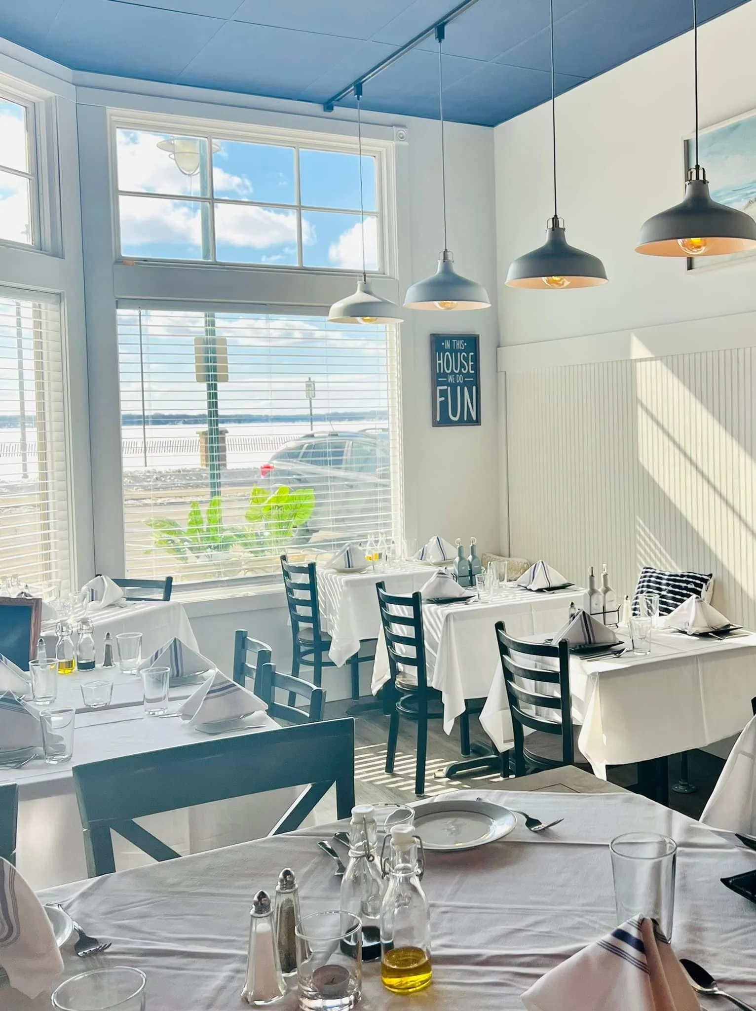 Interior of a bright restaurant with large windows showing a view of a parking lot and water. The tables are set with white tablecloths, napkins, glasses, and condiments. Hanging pendant lights are above the tables, and there's a framed sign on the w