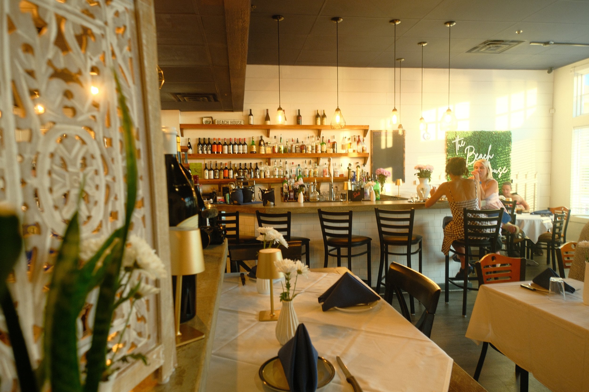 Interior of a bright restaurant or cafe with a bar area on the back wall lined with bottles of alcohol, hanging pendant lights, tables with floral centerpieces, and a few customers seated and chatting.