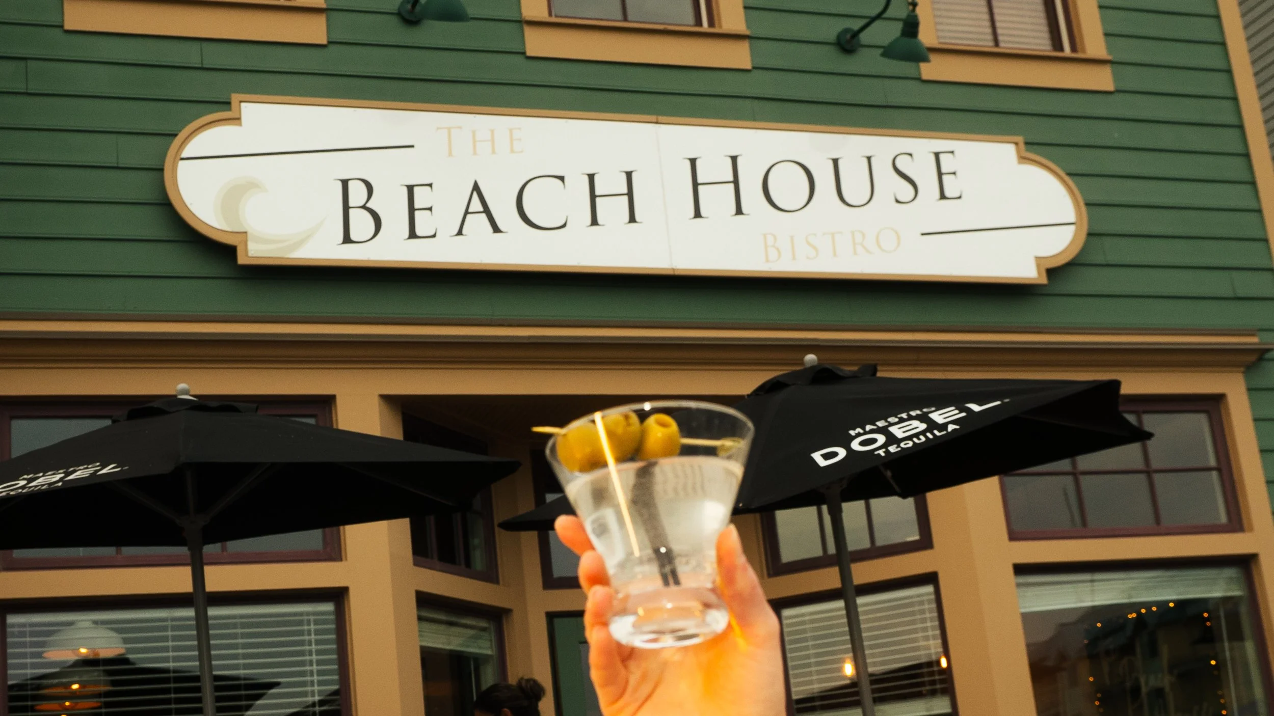 Person holding a cocktail with olives in front of a sign for The Beach House Bistro, with umbrellas and a building in the background.