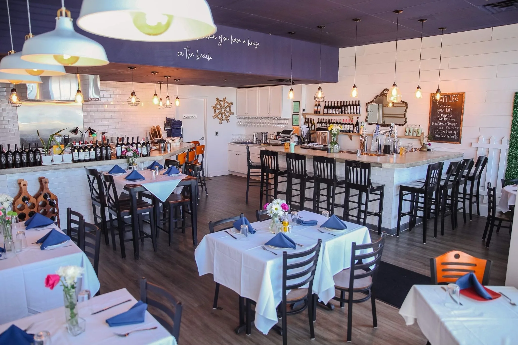 Interior of a restaurant with tables set with white tablecloths, blue napkins, and small flower arrangements. The space features a bar with black chairs, a chalkboard menu on the wall, hanging light fixtures, and a white tiled wall.