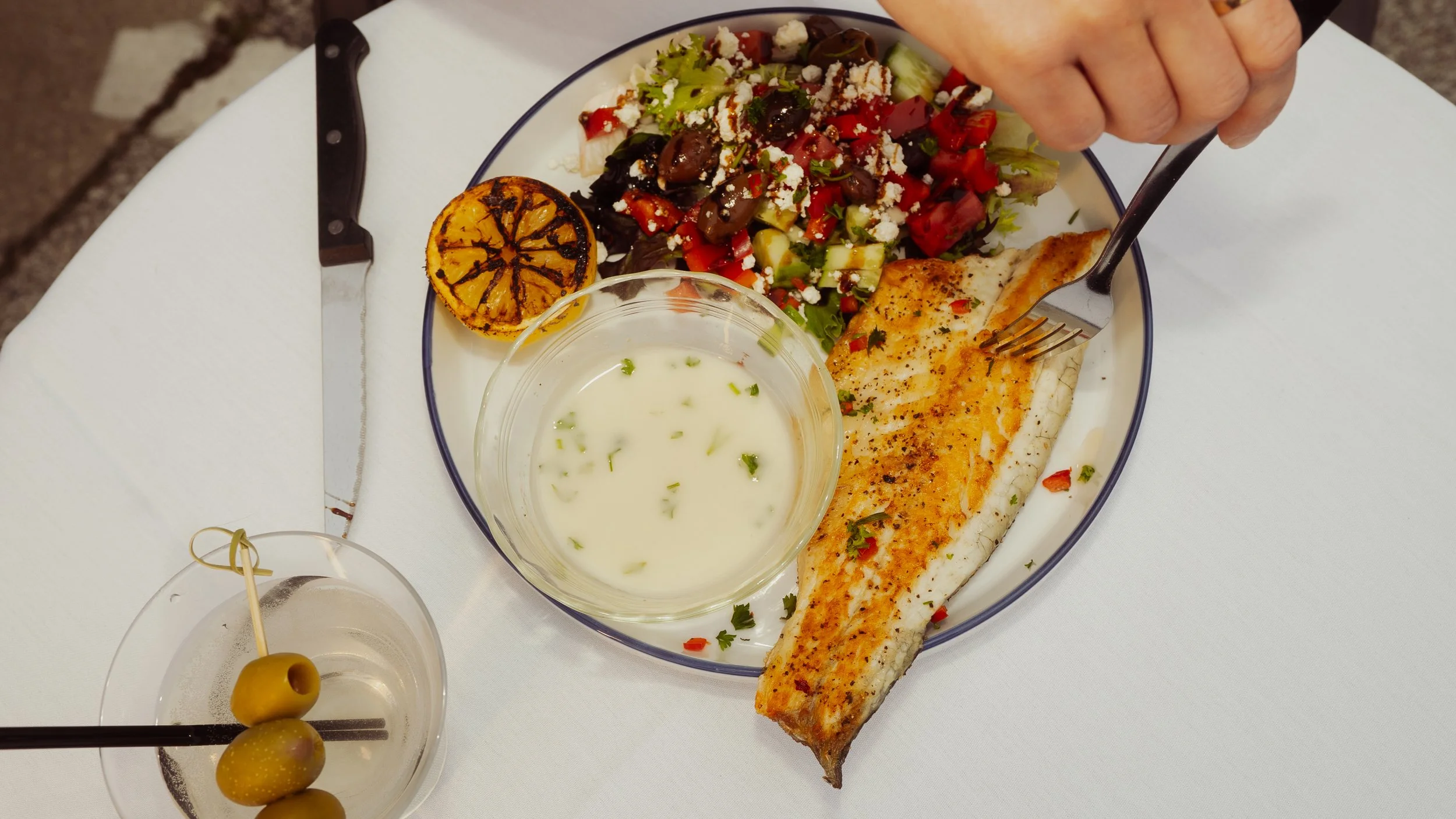 Plate of grilled fish with seasoning, mixed green salad with crumbled cheese, grilled lemon half, small bowl of creamy white sauce, and a glass of drink with green olives on a stick.
