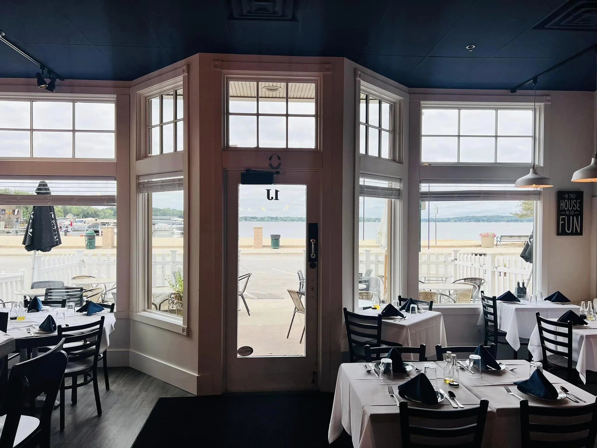 Interior view of a restaurant with tables set for dining, large front windows showing an outdoor view of a marina and water, and a door leading outside.