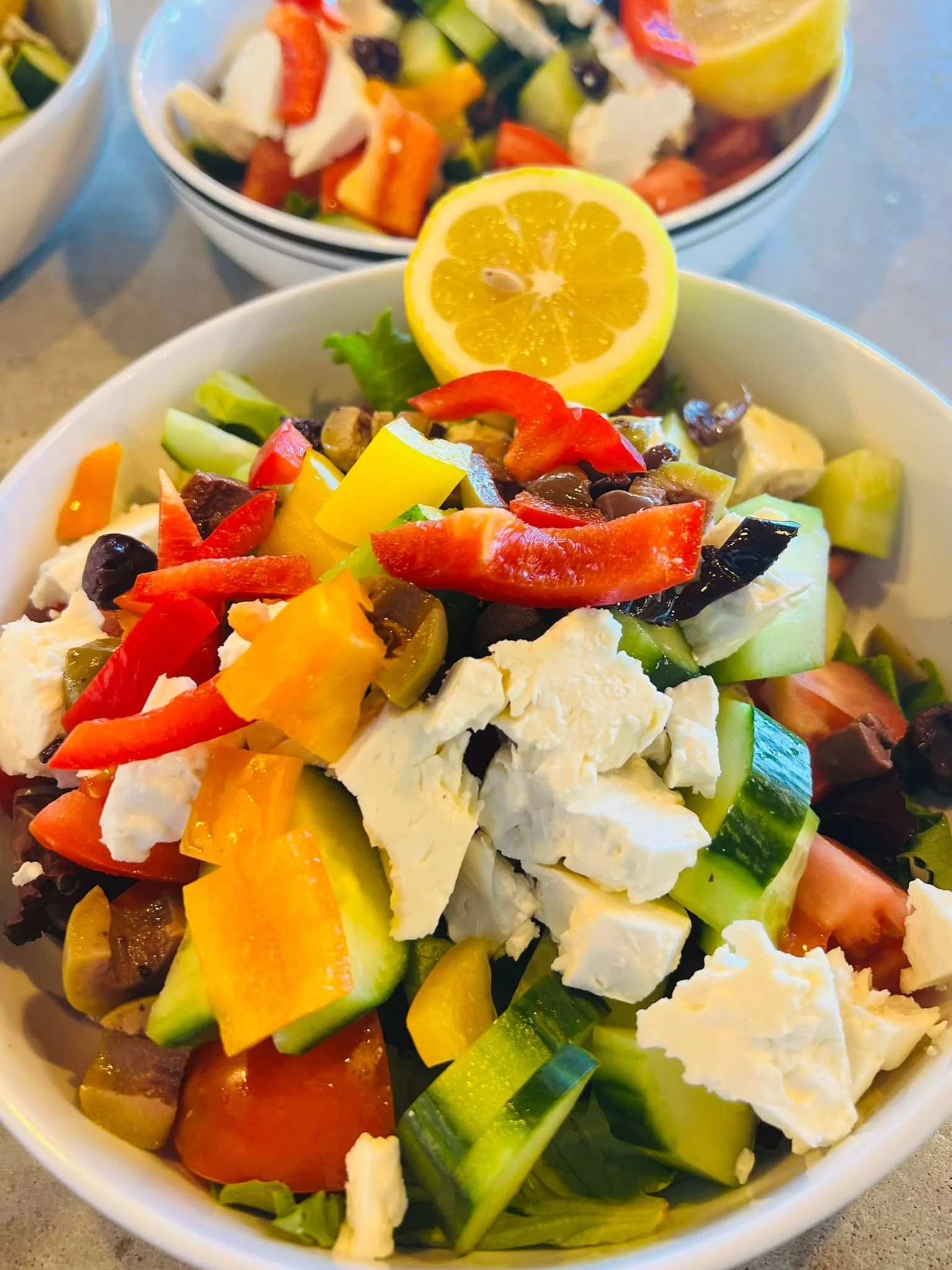 Close-up of a bowl of colorful vegetable salad with chopped tomatoes, cucumbers, bell peppers, olives, and chunks of mozzarella cheese, garnished with a lemon half.