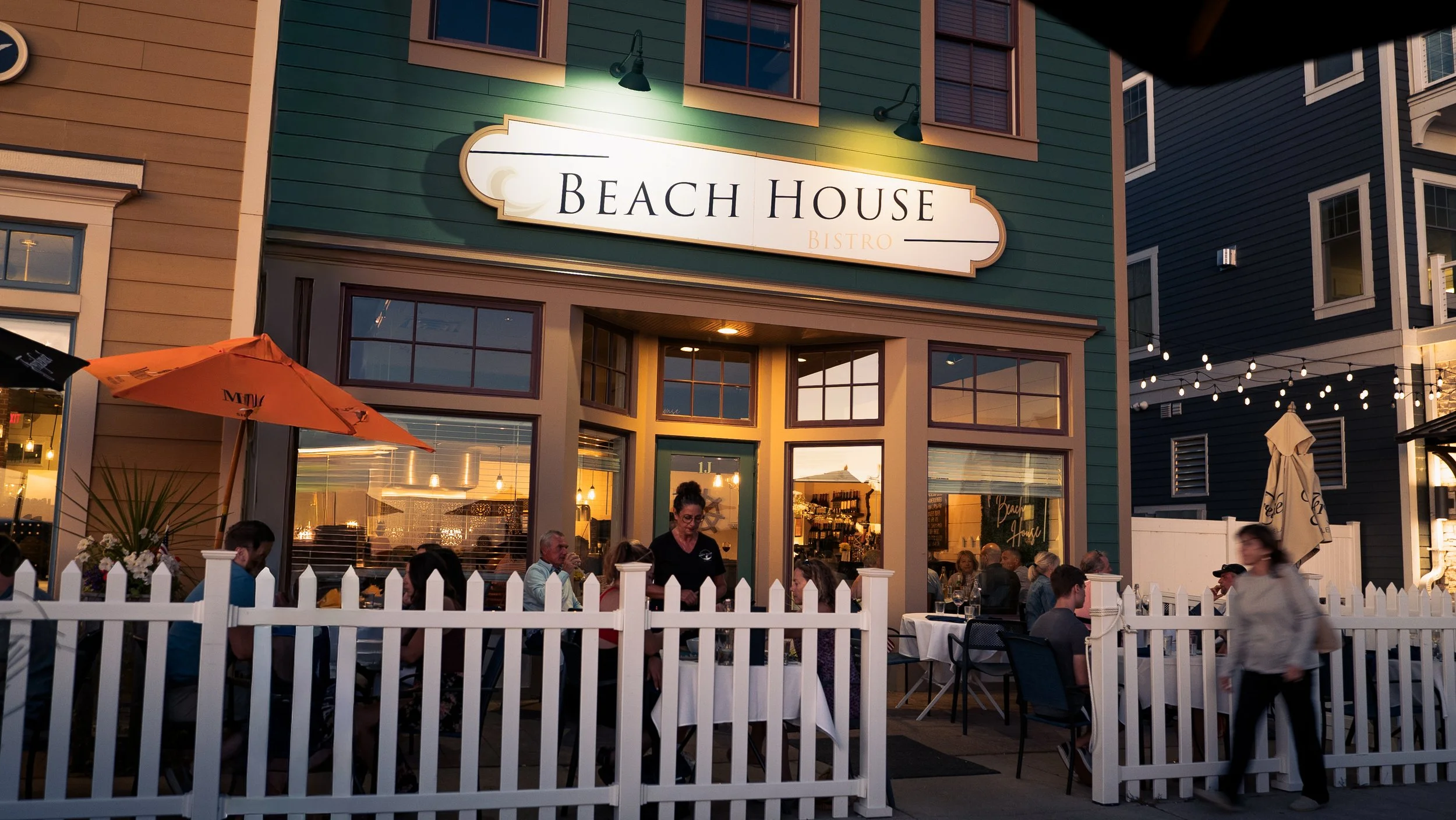 Exterior view of Beach House Bistro with outdoor seating area, people dining, and string lights at dusk.
