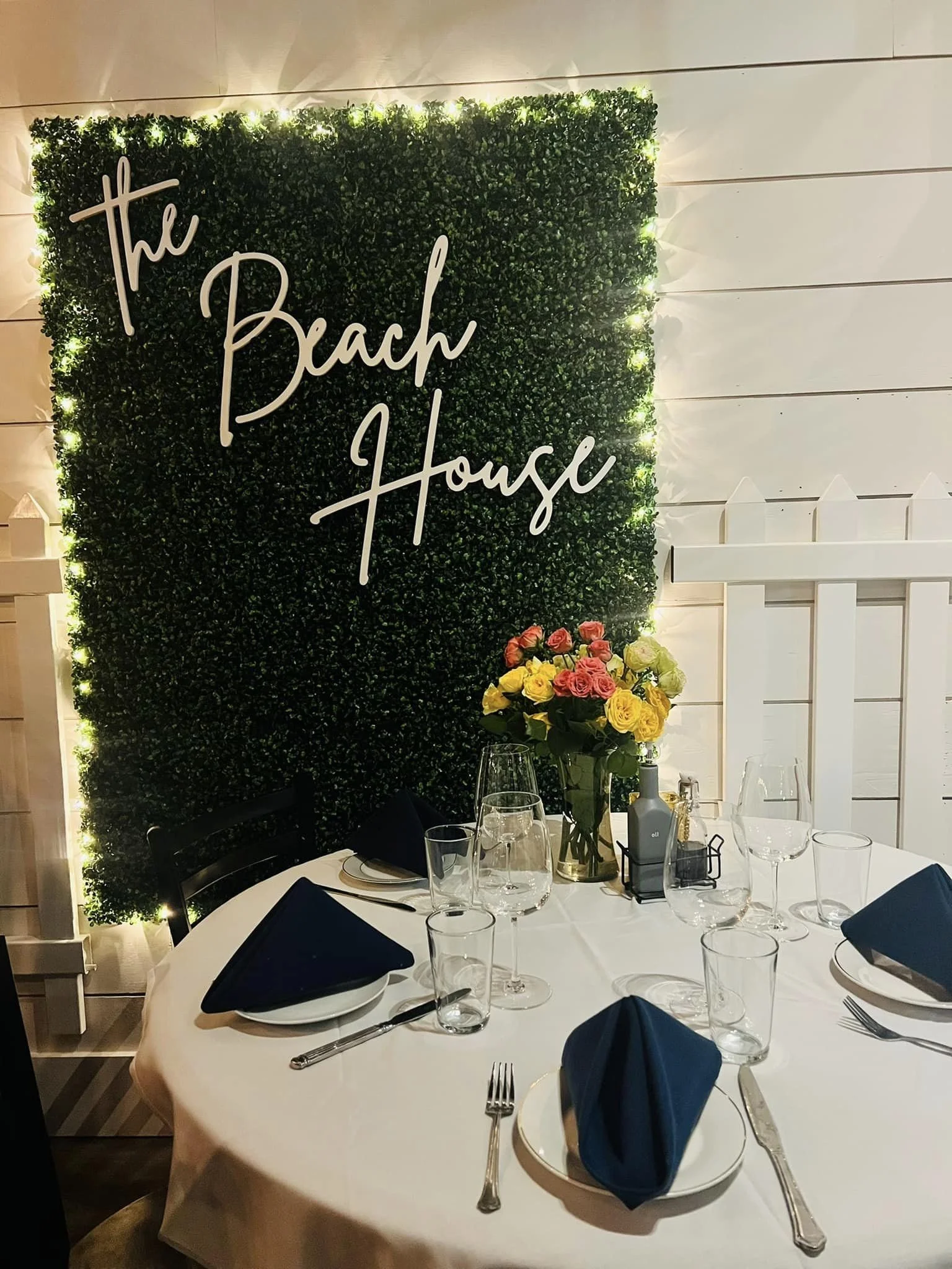 A decorated dining table with black napkins in white plates, set with silverware and drinking glasses. A floral centerpiece with pink and yellow roses sits on the table. A large green hedge wall with illuminated lights and a sign reading 'The Beach H