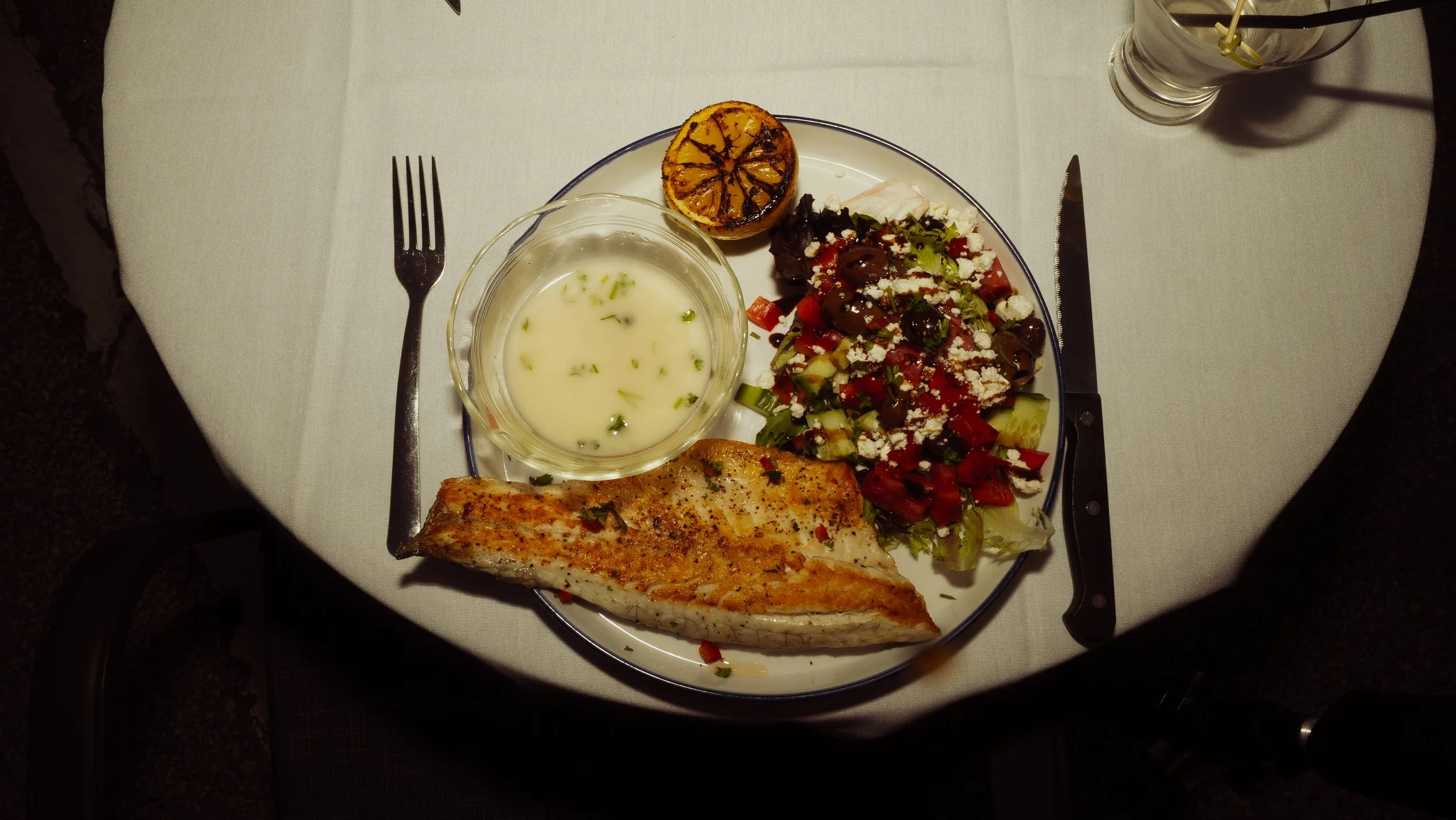 A plate with grilled fish, a mixed salad with cheese, a bowl of creamy dressing, and a grilled lemon half on a white tablecloth, with a water glass and utensils.