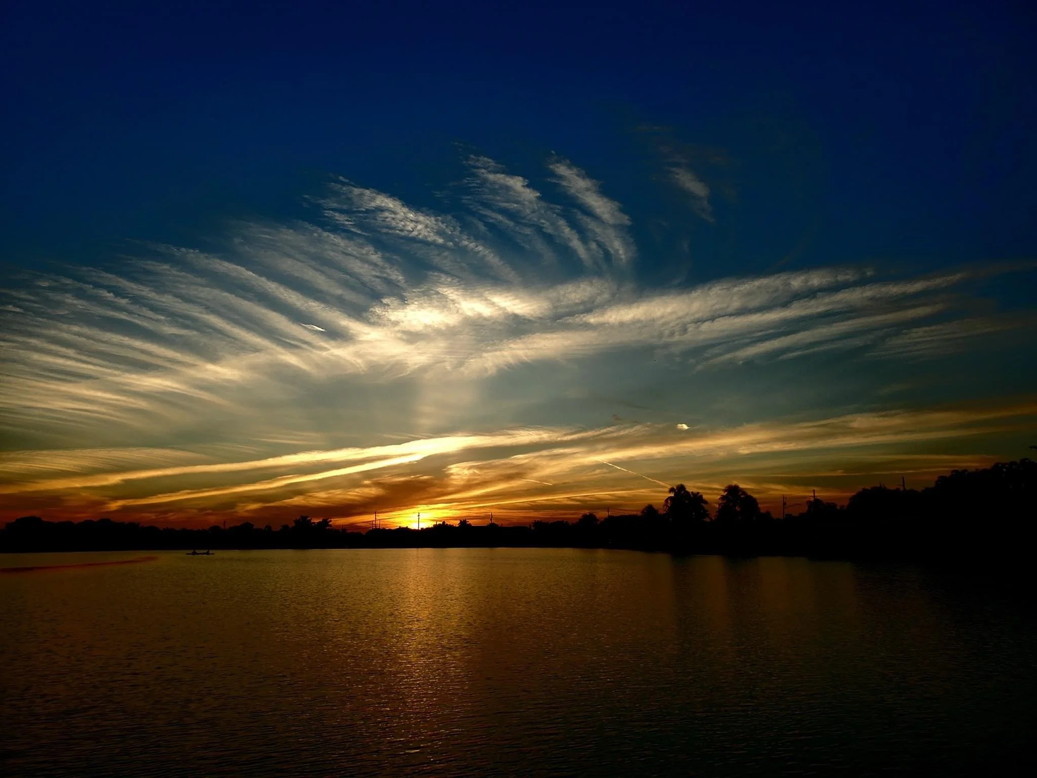A sunset over a body of water, with a dark silhouette of trees and the sky filled with scattered, wispy clouds illuminated by the setting sun.