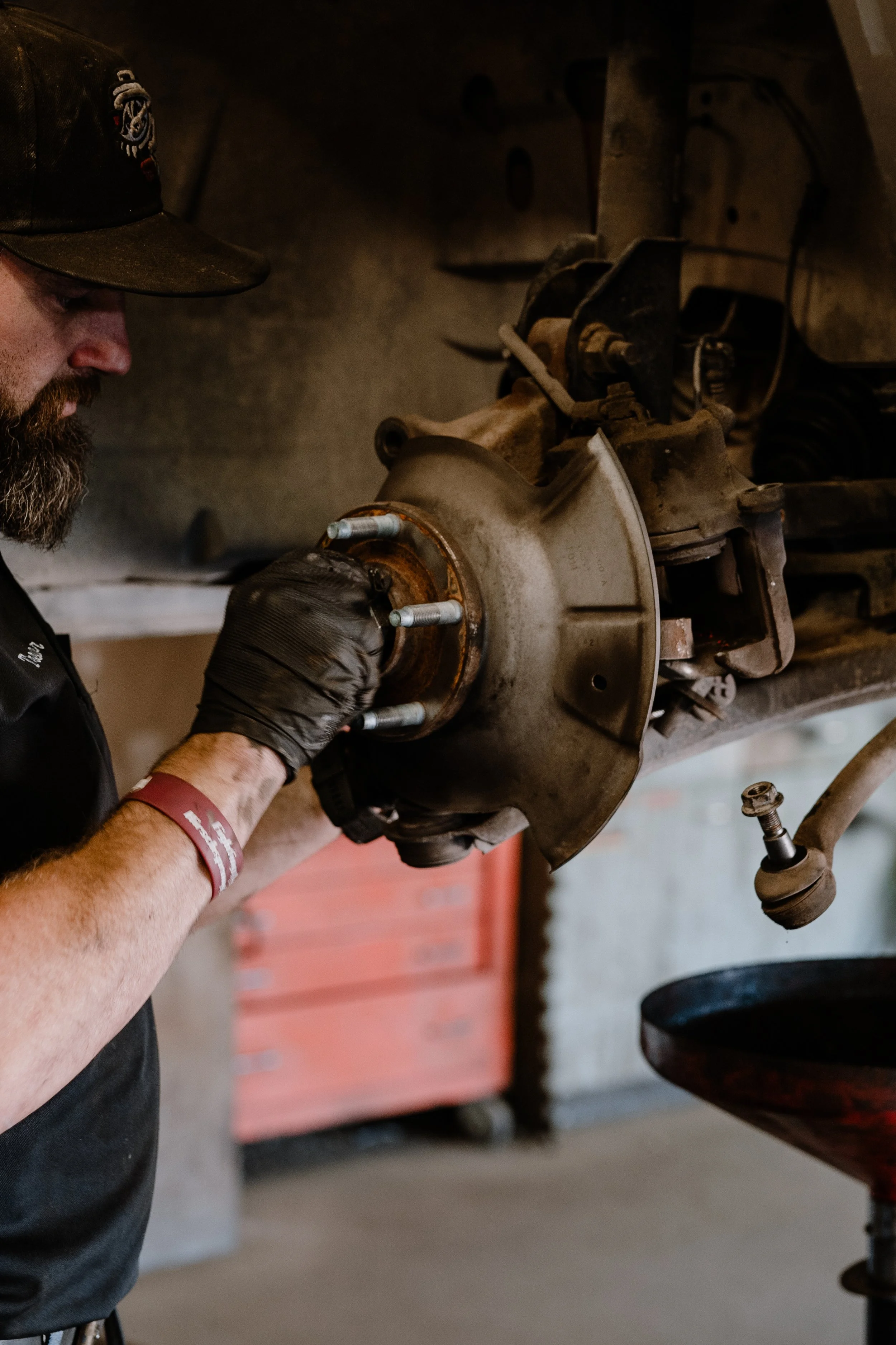 A mechanic working on a vehicle's brake assembly, holding a tool and wearing gloves in a garage.