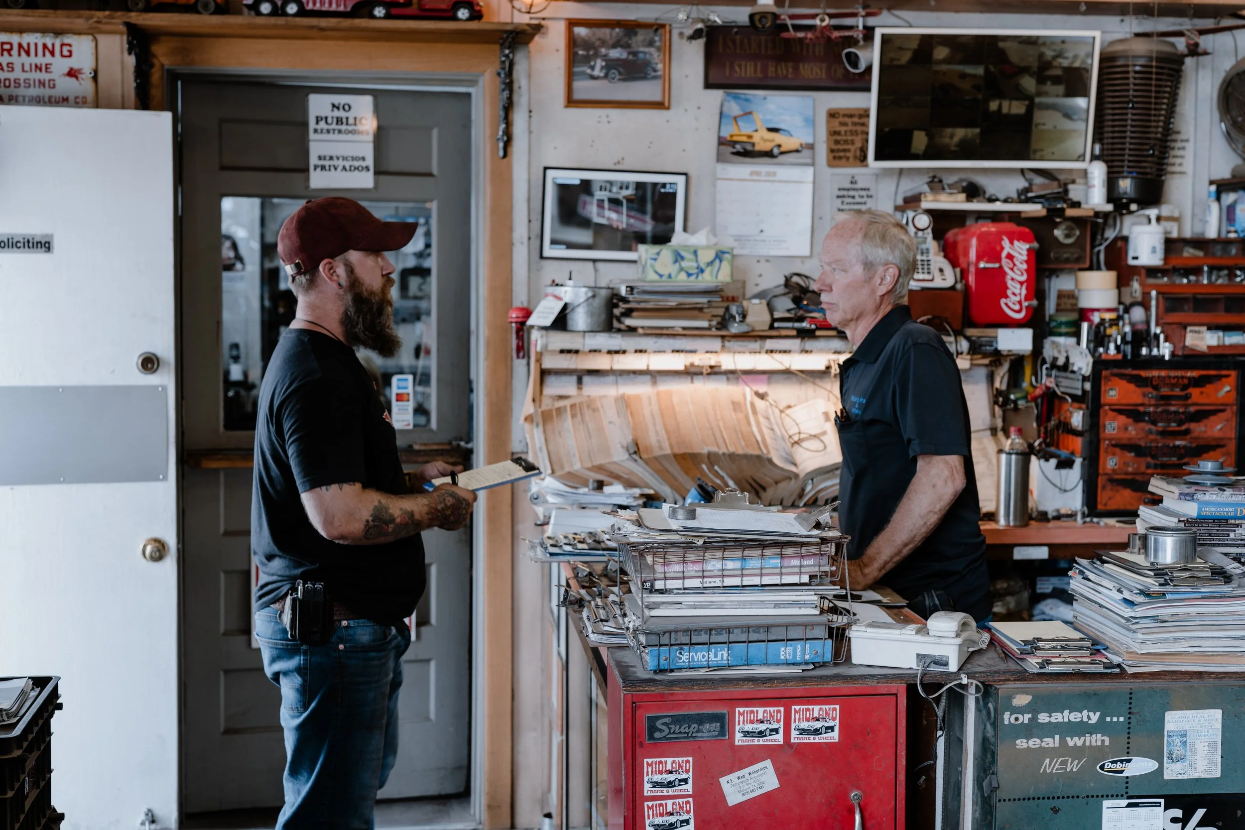 Two men face each other and are engaged in a conversation inside a cluttered auto repair shop or workshop, with tools, stereo equipment, papers, and framed pictures on the wall behind them.