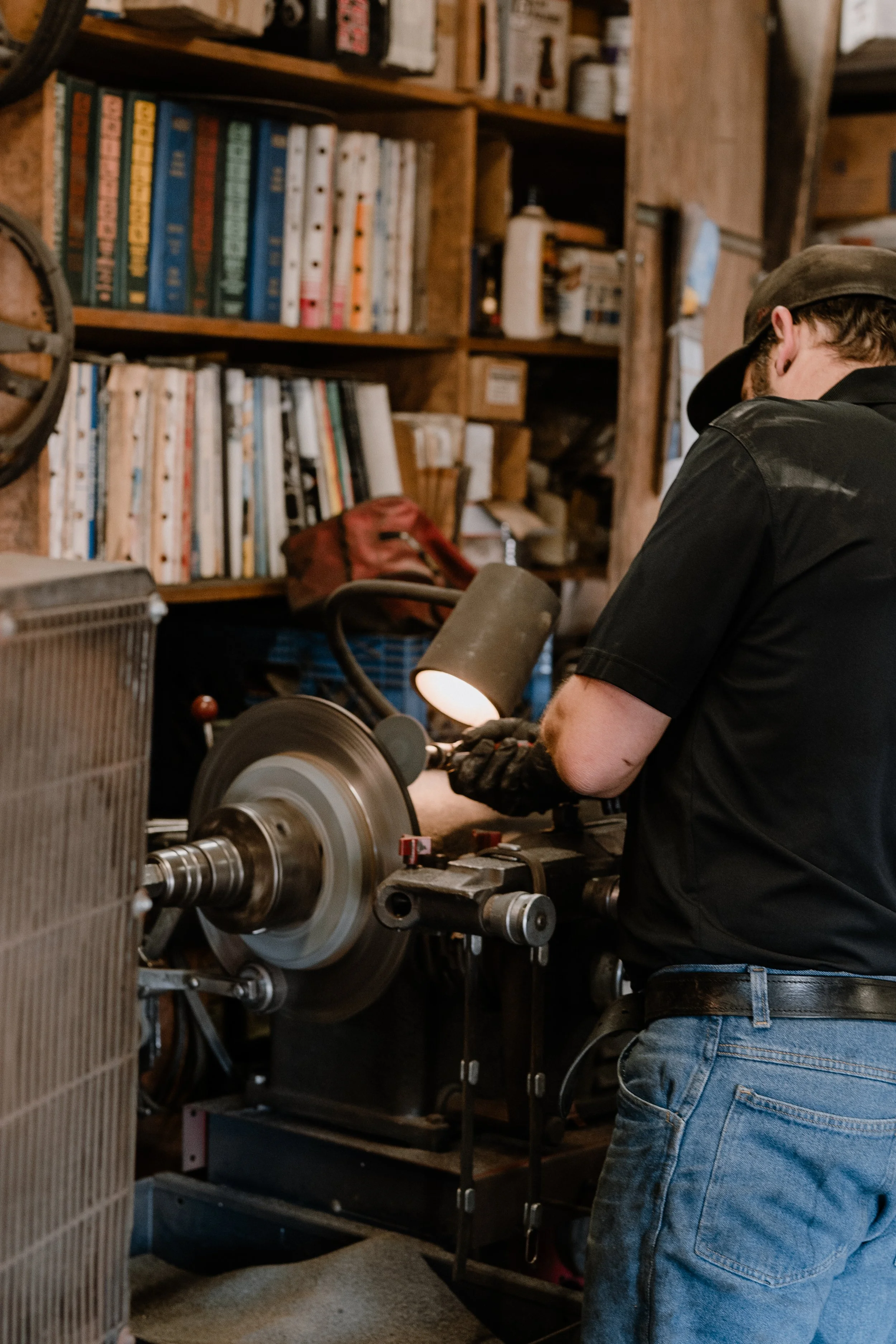 A person in jeans and a black shirt working at a metal lathe in a workshop with wooden shelves filled with books and supplies in the background.