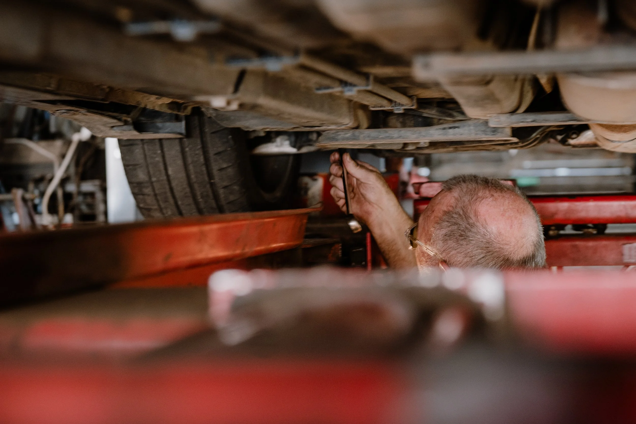 A person working underneath a vehicle, using a wrench, with a tray positioned underneath the vehicle, in a garage or workshop setting.