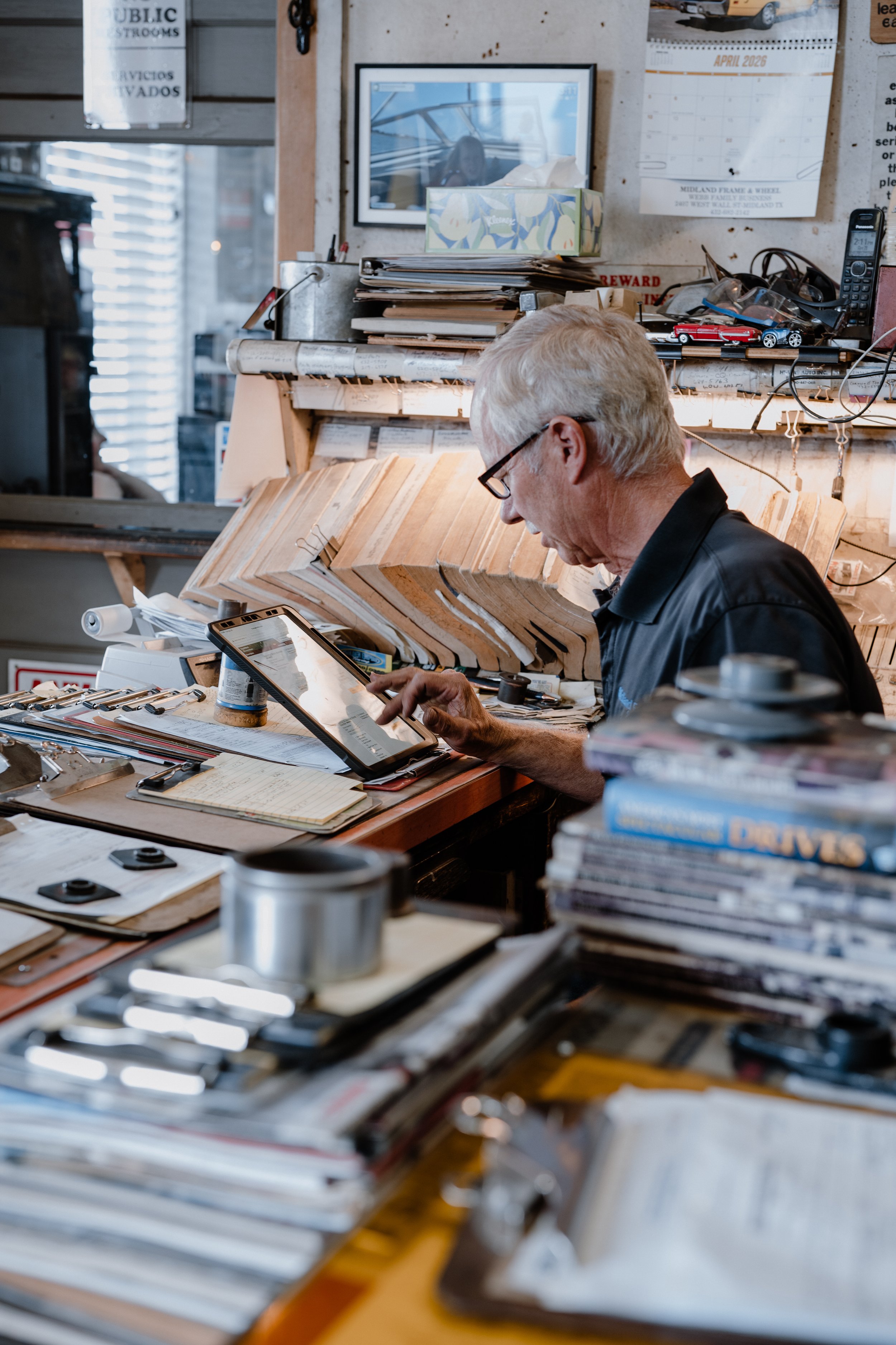 A man with gray hair and glasses working at a cluttered desk filled with papers, books, and office supplies in a busy workspace. There are framed pictures, calendars, and various objects on the wall behind him.