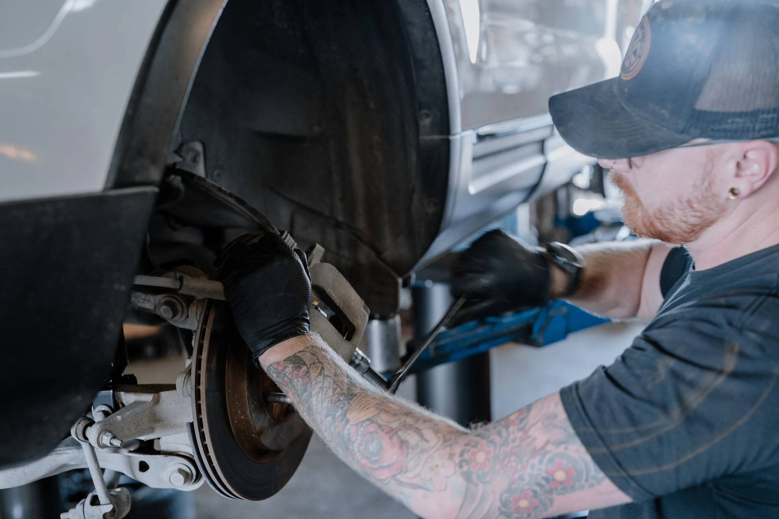 A tattooed person wearing a cap and glasses working on a car's brake system in a garage.