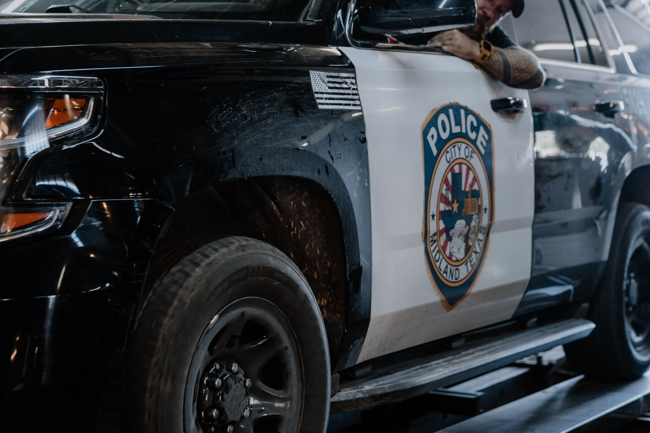 A police vehicle with a partial badge and an American flag decal on the side, parked in a parking lot. A person with a tattooed arm, wearing a hat and a watch, is leaning into the vehicle.
