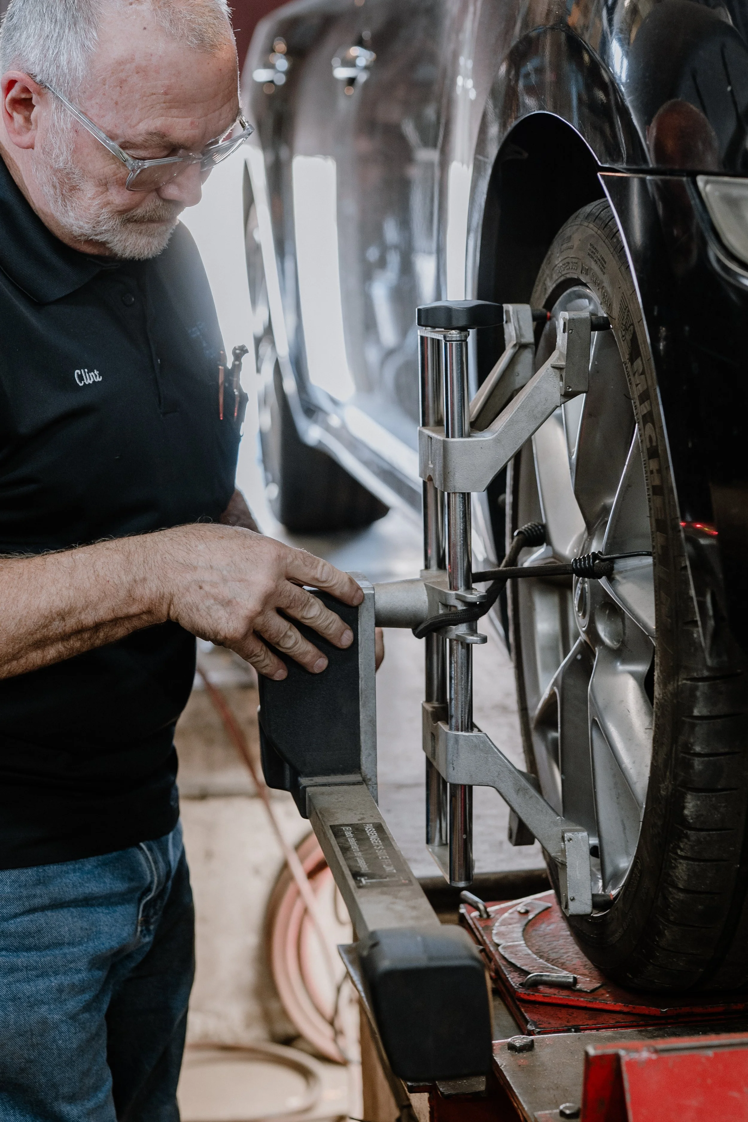 A man wearing safety glasses and a black shirt labeled "Clint" is working on a car tire using a machine in a garage or workshop.
