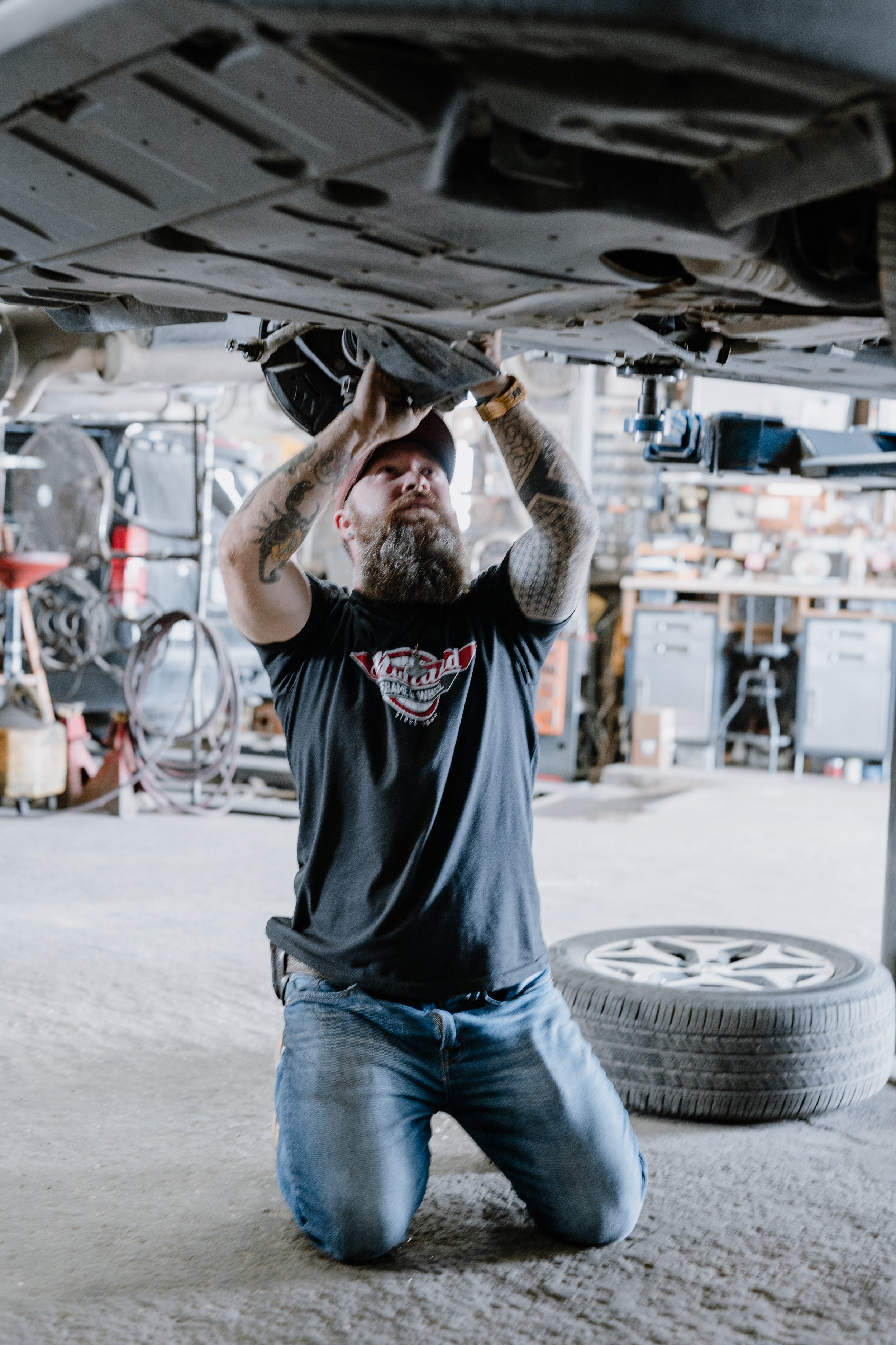 A man with tattoos on his arms and a beard, wearing a black t-shirt and jeans, kneeling on the ground and working under a vehicle in a workshop, with tools and car parts around him.