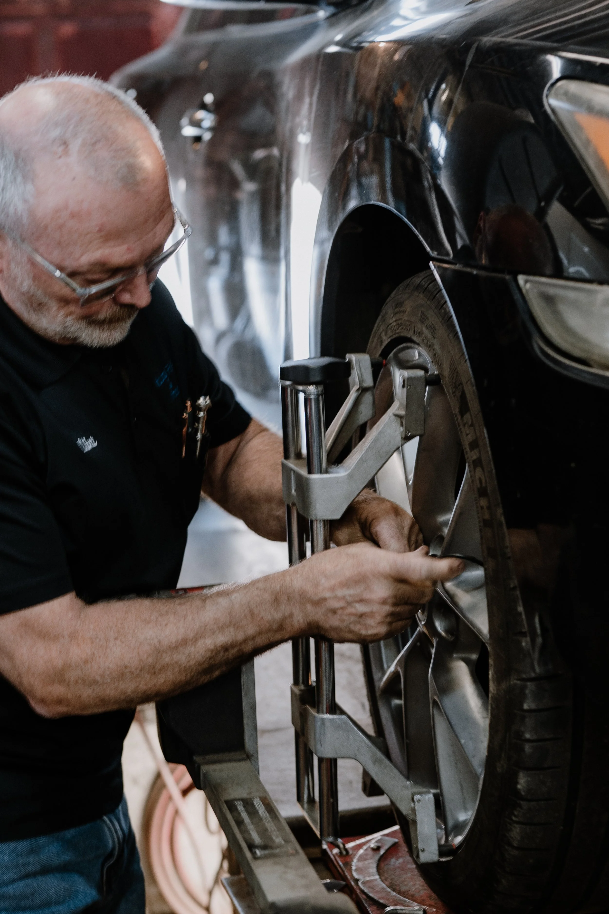 An older man with glasses and a beard is working on a black car using a tool. He is wearing a black shirt and is focused on the wheel area, which is mounted on a repair stand in a garage or workshop.