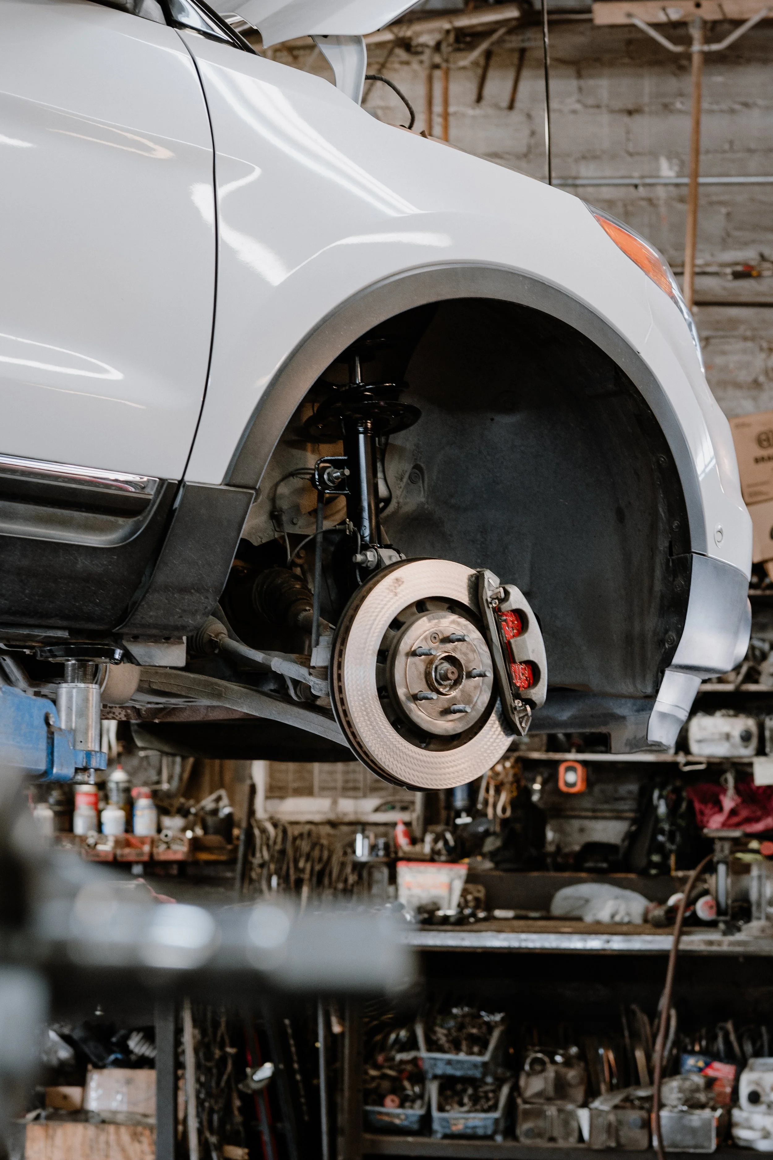 A white car is lifted on a hydraulic lift inside a garage or repair shop, with the wheel removed, exposing the brake disc and suspension components. The background includes various tools and supplies.