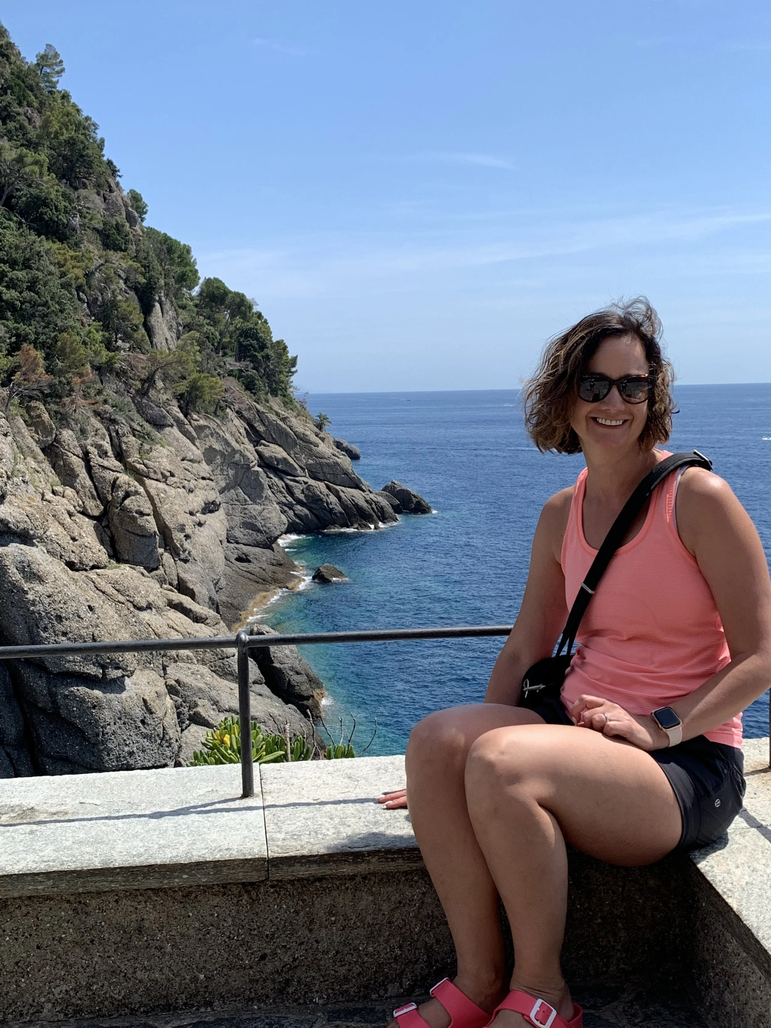 A woman with curly brown hair, wearing sunglasses, a pink tank top, and black shorts, sitting on a stone ledge near the ocean with rocky cliffs and green trees in the background.