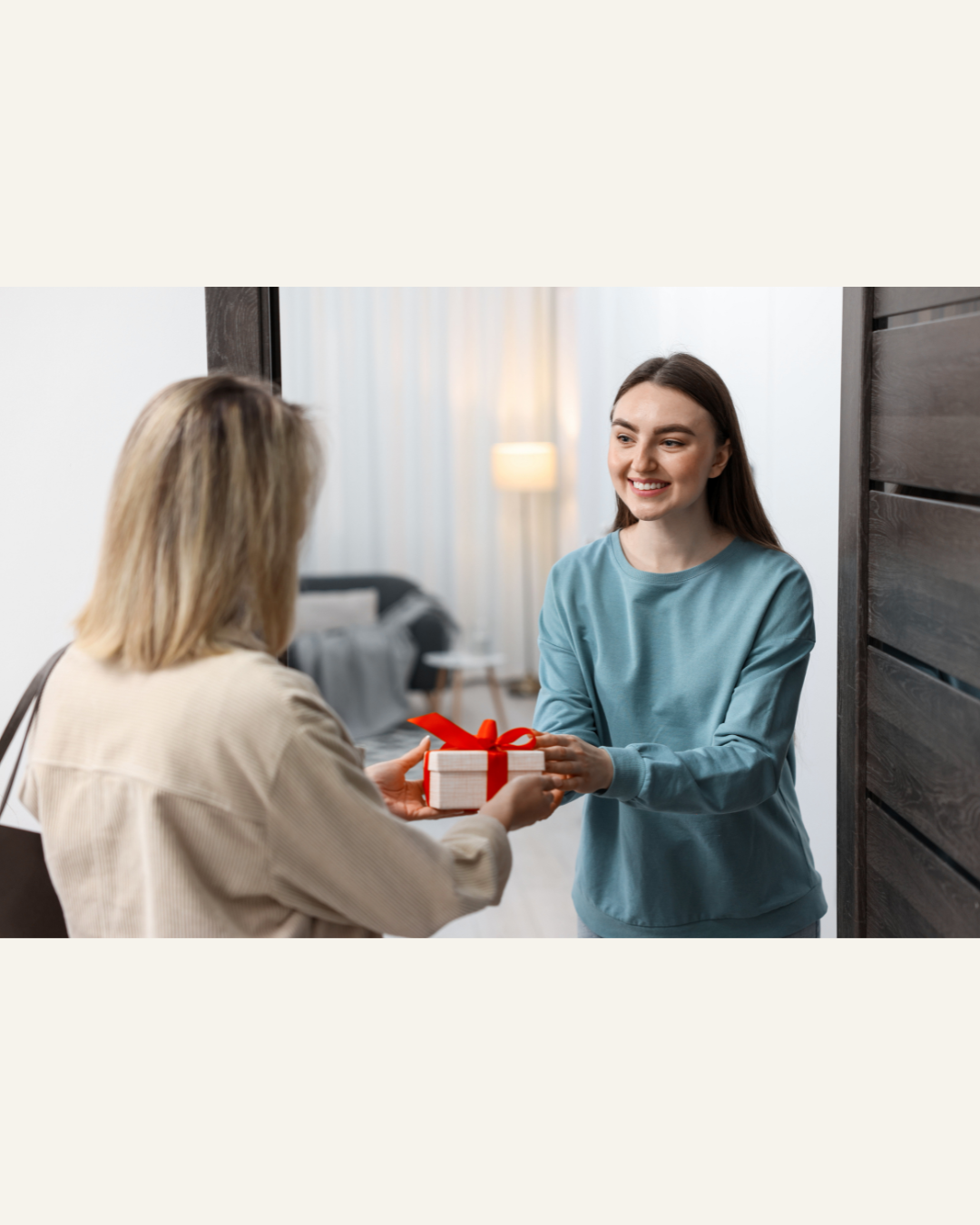 a woman with blonde hair handing a gift with a red ribbon to a younger woman in the entrance of her home
