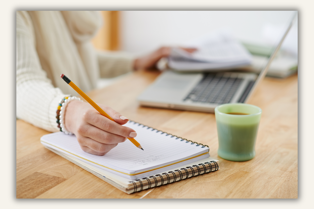 a person wearing cream sweater, a wooden table with a computer open and writing on a notepad