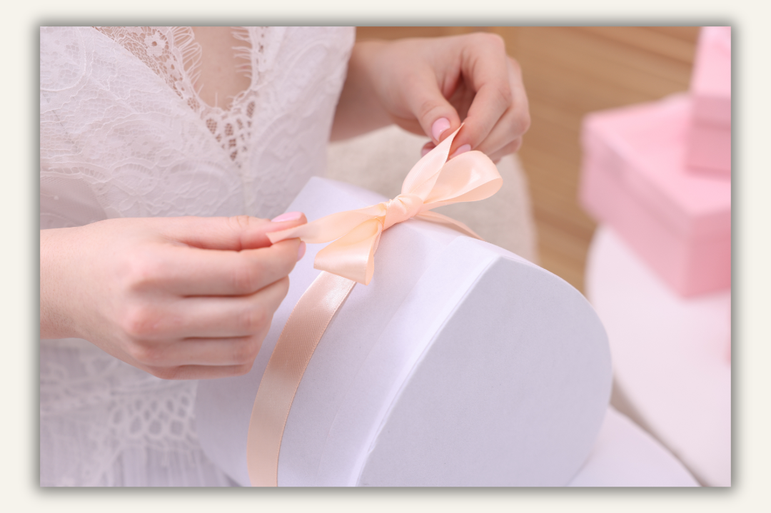 a bride wearing white lace dress open a white heart shaped gift with a peach coloured ribbon, 2 pink gift boxes in the background