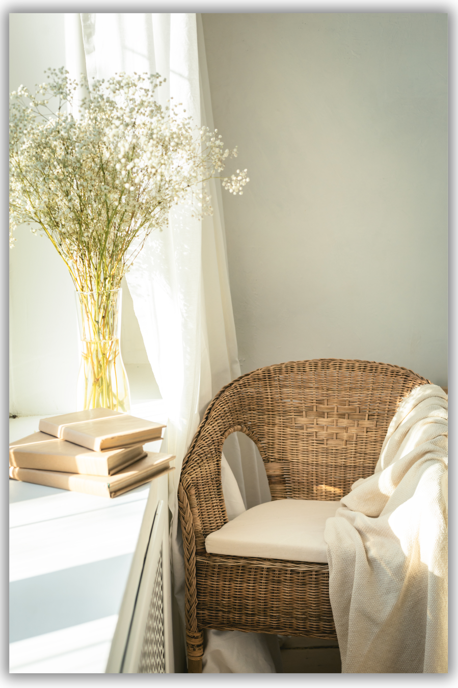 wicker chair with throw blanket on it, white table with 3 brown books stacked along with a clear vase containing baby breath
