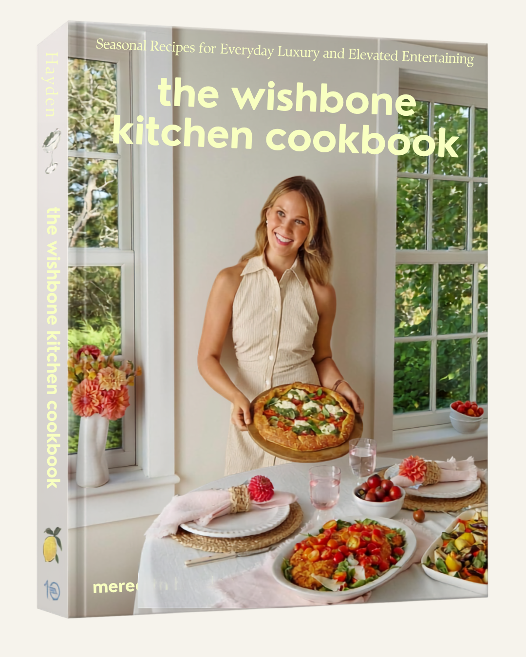 cookbook with title the wishbone kitchen cookbook, a women holding a pizza behind a beautifully decorated table with food on it