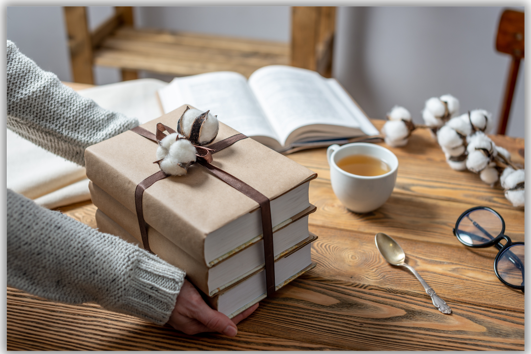3 books wrapped in brown paper with ribbon, cup of tea, spoon and open book on a wooden table