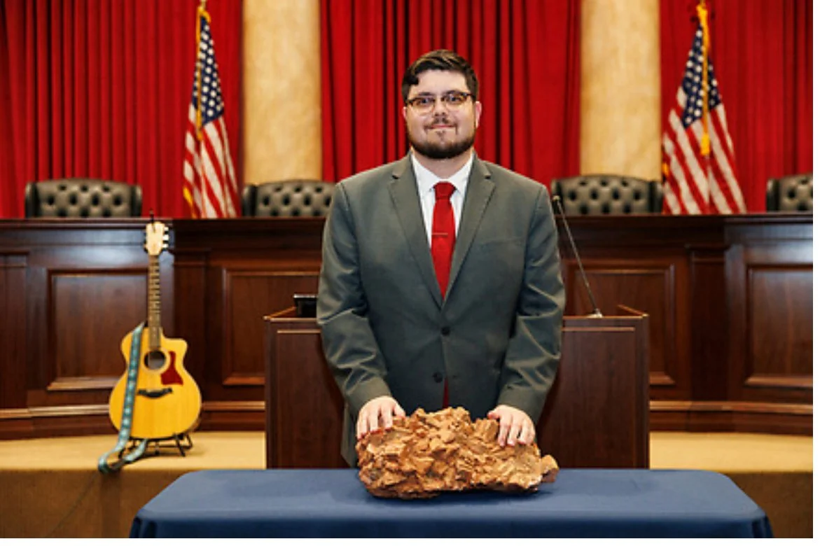 A man in a suit standing behind a table with a large rock, in a courtroom setting with American flags and red curtains in the background.