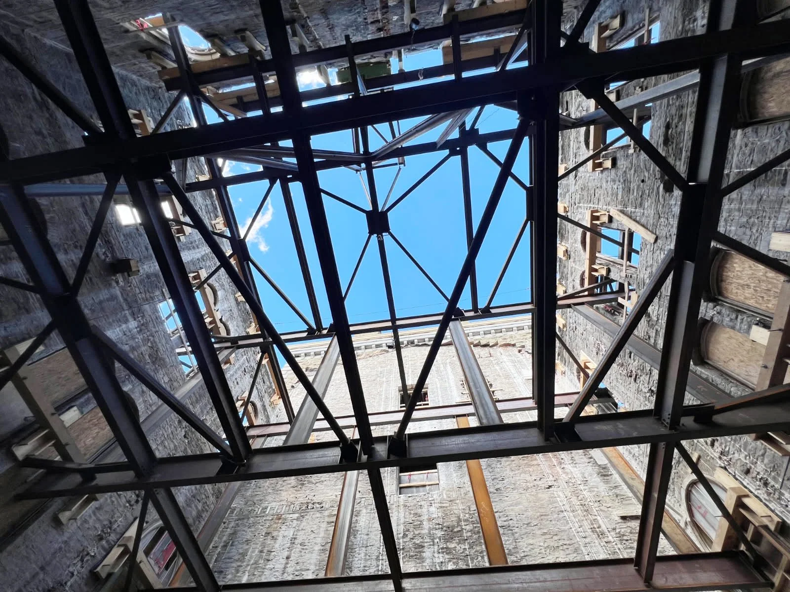 Looking up into a building under construction, showing a square opening at the top with sky visible, surrounded by scaffolding and construction materials.